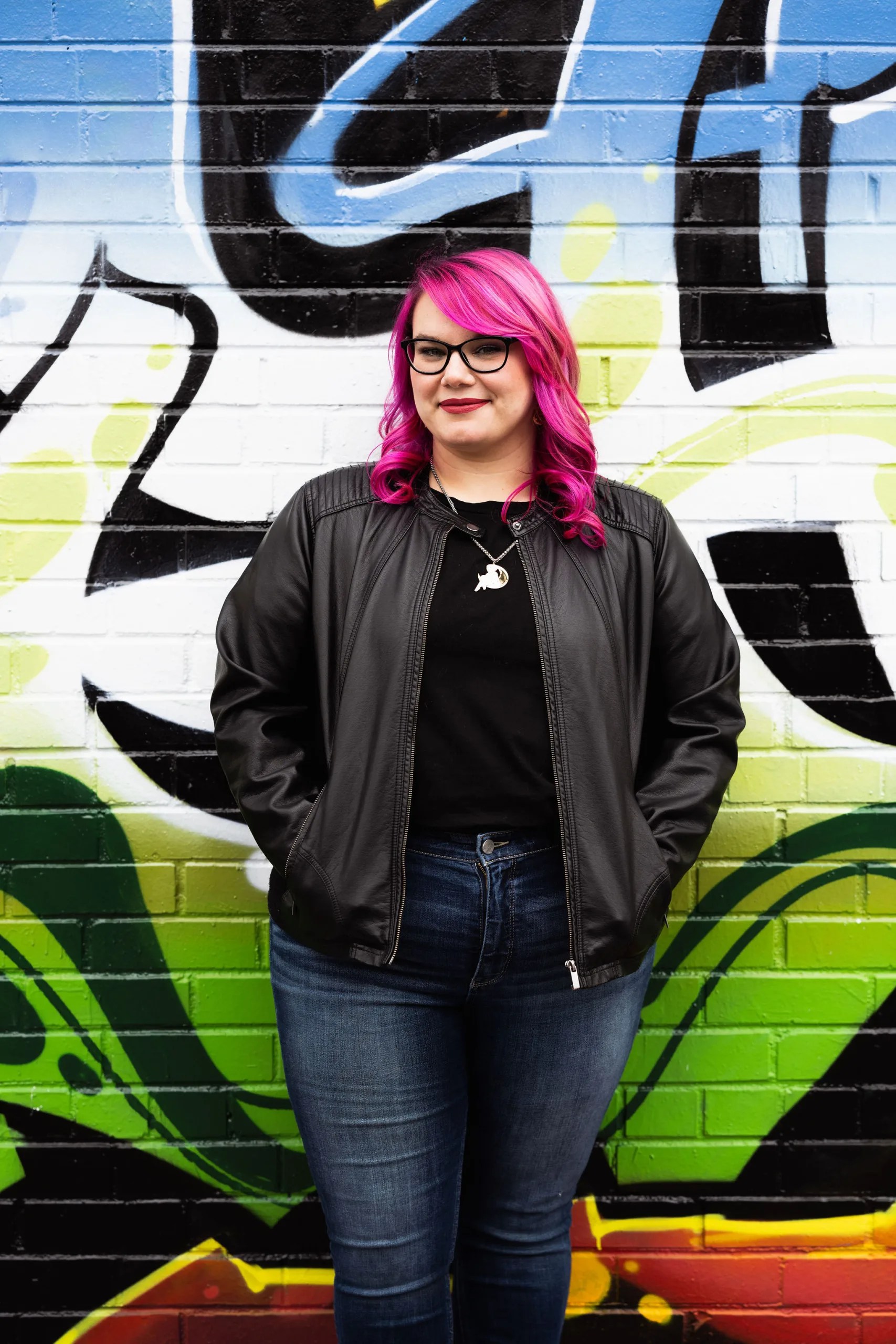 female with bright prink hair wearing a black leather jacket and jeans in front of a graffitied wall