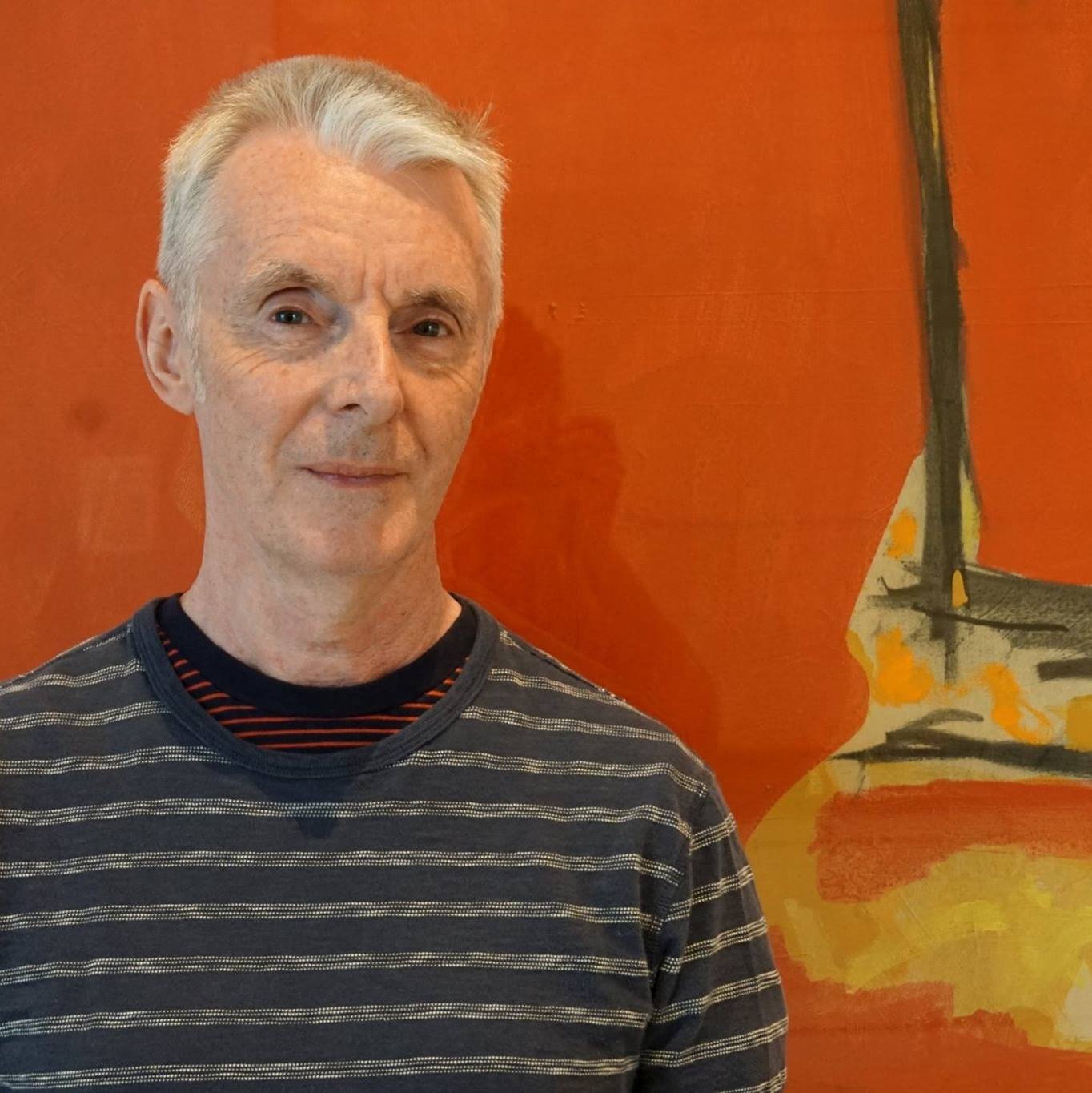 Man with short grey hair wearing a navy and white striped shirt standing in front of a piece of artwork which shows orange yellow and black colors