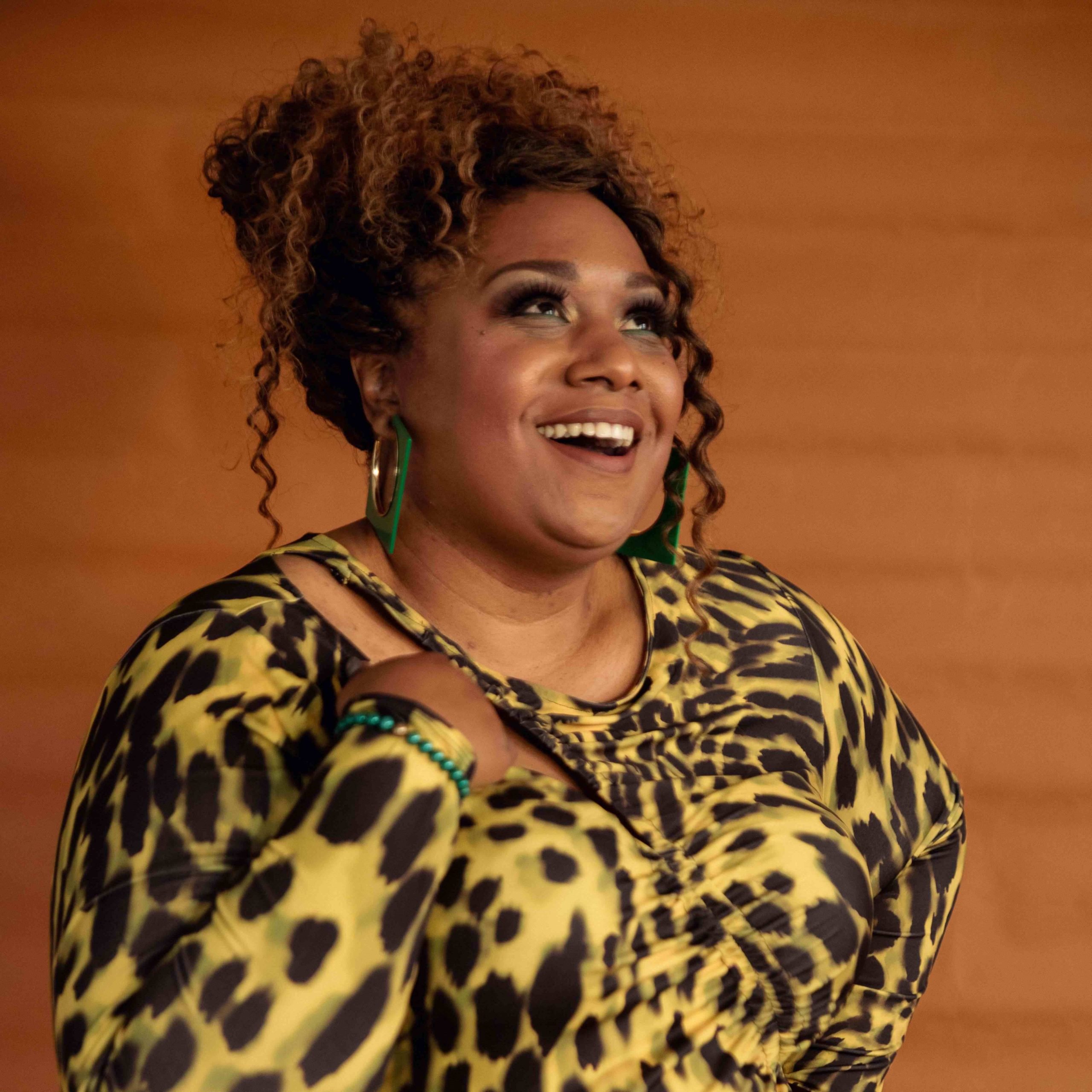 woman with curly hair smiling wearing a leopard print top looking towards the top right of the frame