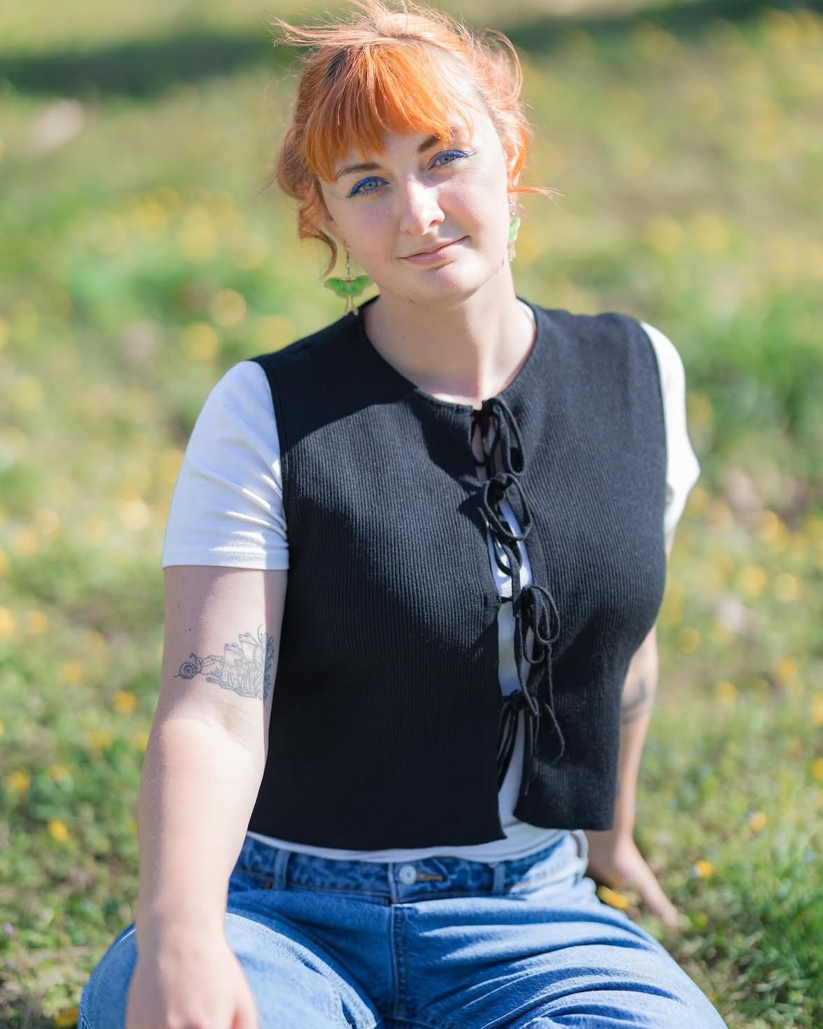 a person with orange red hair wearing a black sweater vest and blue jeans sitting in grass