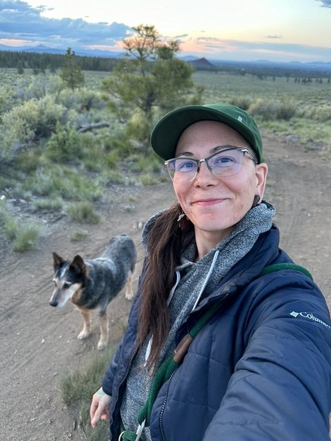 smiling woman in a grey jacket and grey cap wearing glasses in front of an outside background with a black haired dog