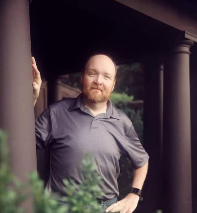 bald man with short beard and moustache in a grey polo style shirt leaning on a column with his hand on his hip