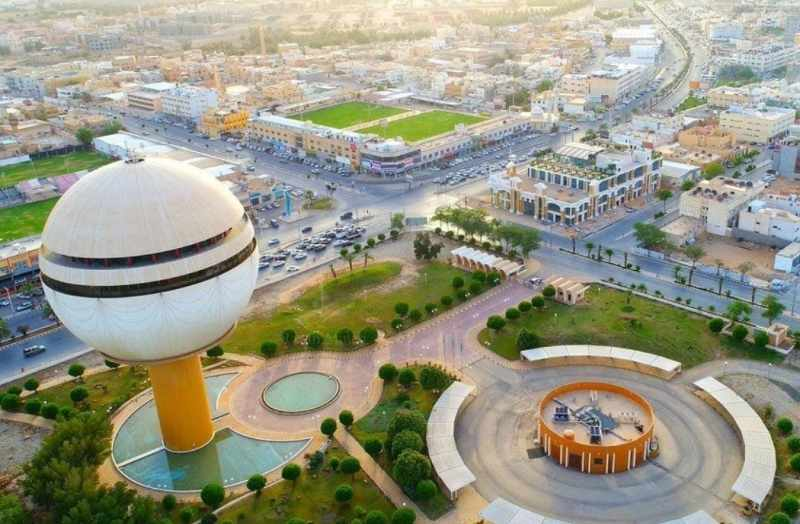 منظر جوي لمنطقة حضرية تضم برج مياه بارز ومساحات عامة خضراء ومبانٍ سكنية وتجارية.