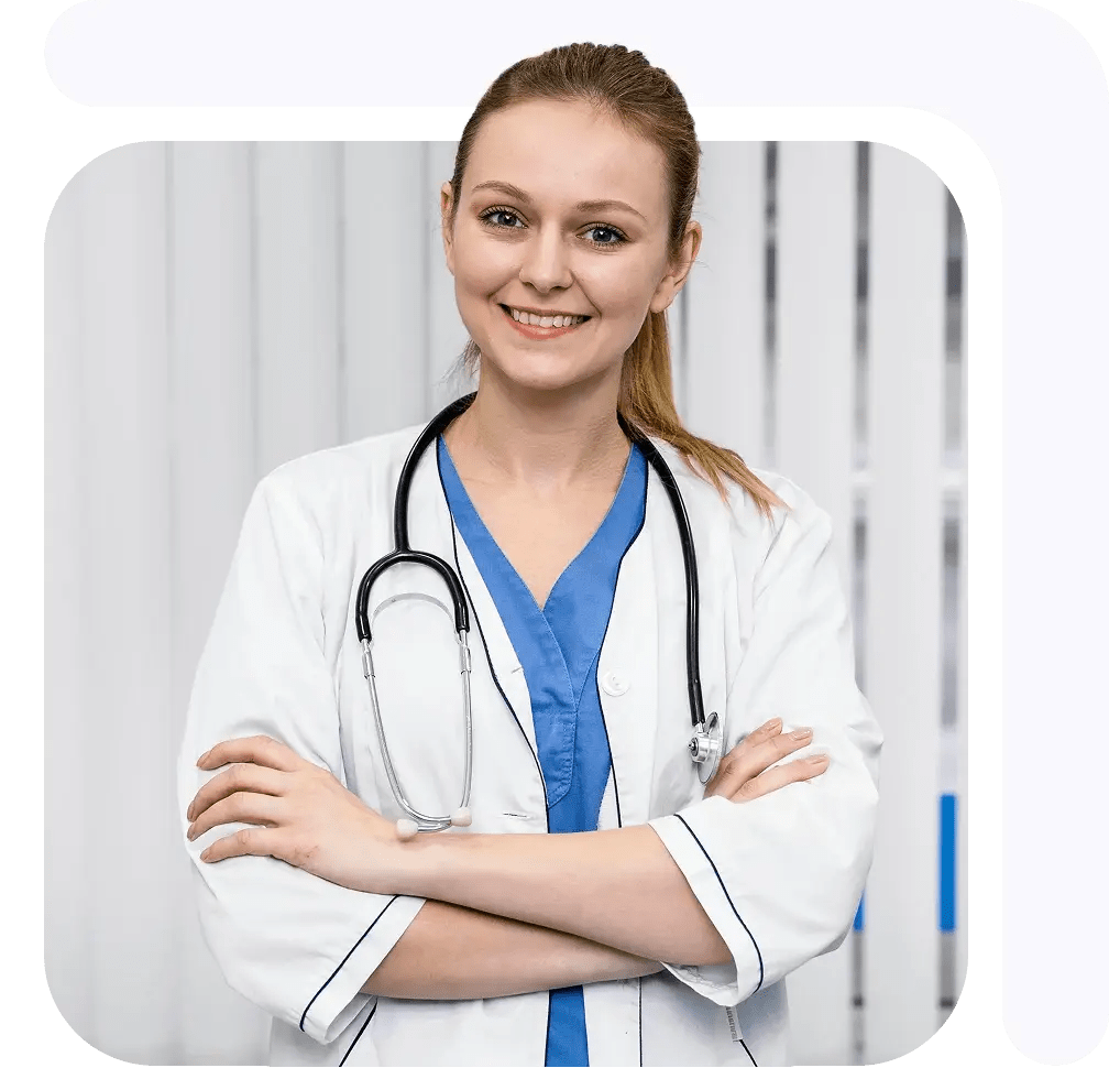 Female medical professional in a white coat with a stethoscope, smiling and standing with arms crossed.
