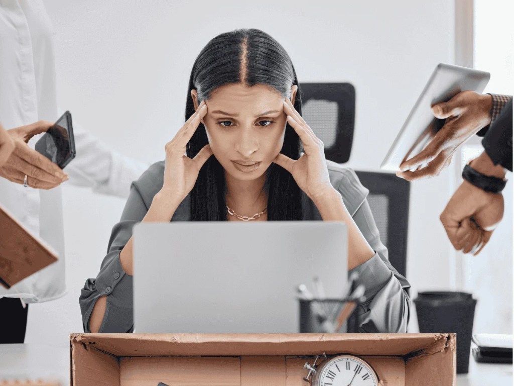 Overwhelmed woman holding her head at a laptop while surrounded by people with devices and papers