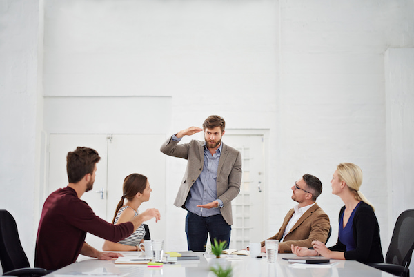 A-picture-of-three-male-employees-and-two-female-employees-having-a-brainstorming-collaborative-session-one-of-the-males-is-standing-at-the-front-of-the-other-sharing-his-ideas