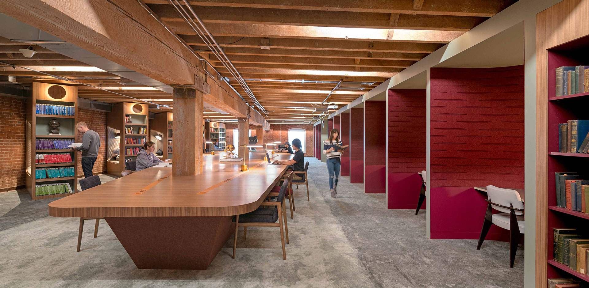 Intérieur d'une grande bibliothèque moderne au design industriel et chaleureux. Le plafond présente de massives poutres en bois brut. Une immense table centrale en bois verni s'étend sur toute la longueur de la pièce, équipée de lampes de lecture minimalistes dorées où plusieurs ingénieurs travaillent sur des ordinateurs portables. À droite, une rangée d'alcôves de lecture individuelles est tapissée d'un revêtement acoustique rouge bordeaux. À gauche, de grandes étagères de bibliothèque encadrent des murs en briques rouges.