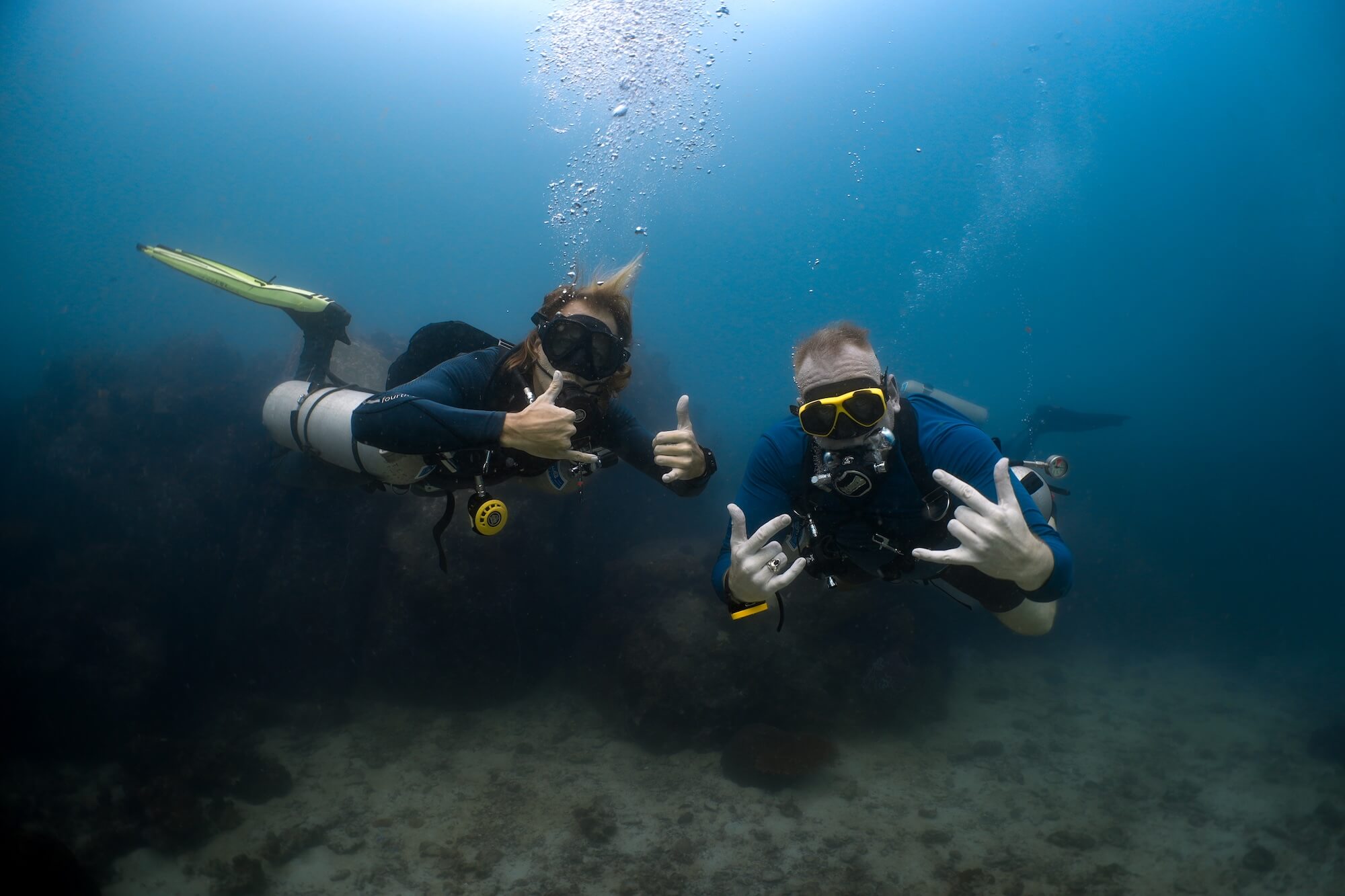 Two scuba divers underwater giving thumbs-up and hand signals, surrounded by coral reef in clear ocean water.
