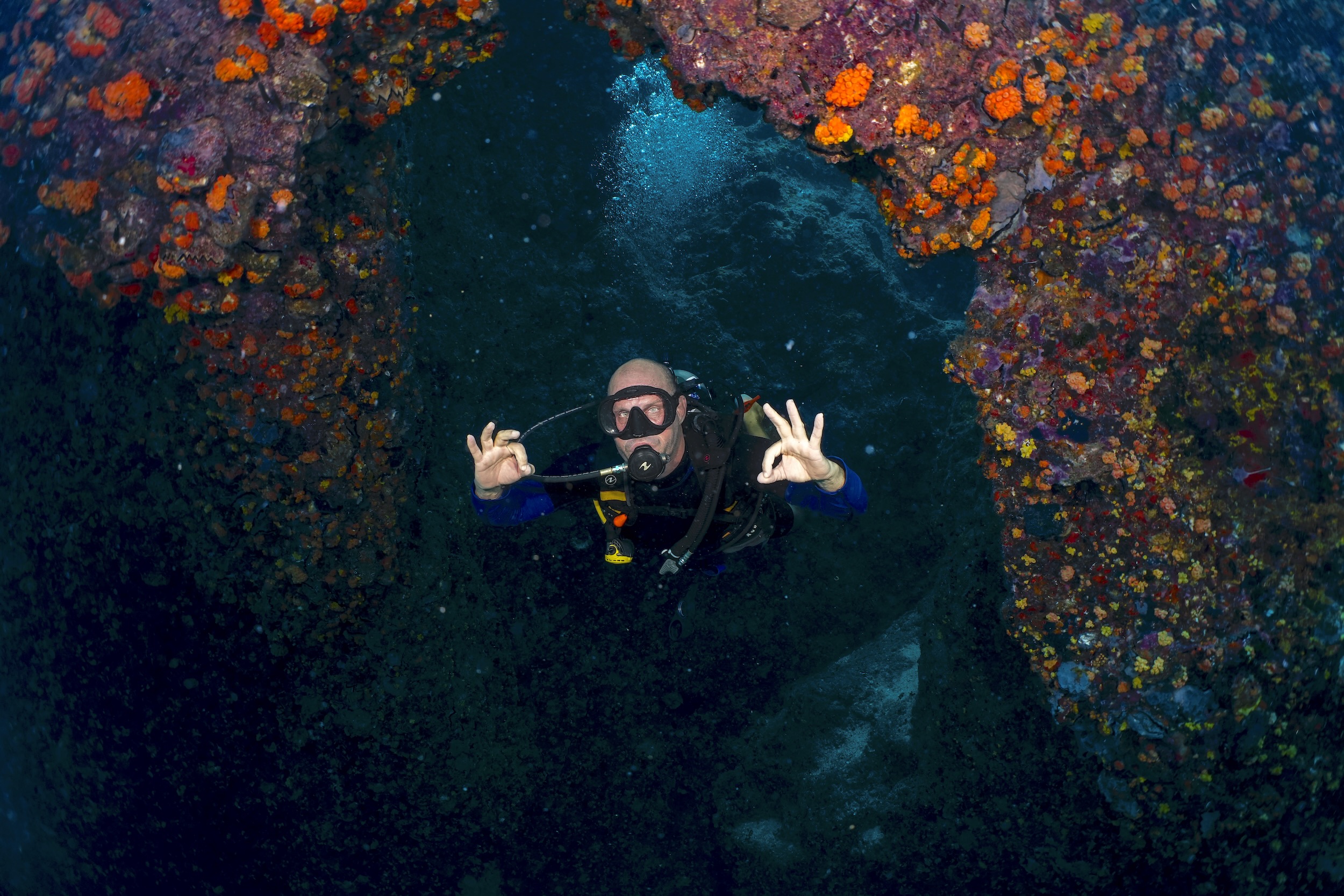 Diver exiting the Chimney swimthrough at Sail Rock, making OK sign, surrounded by colorful marine life.