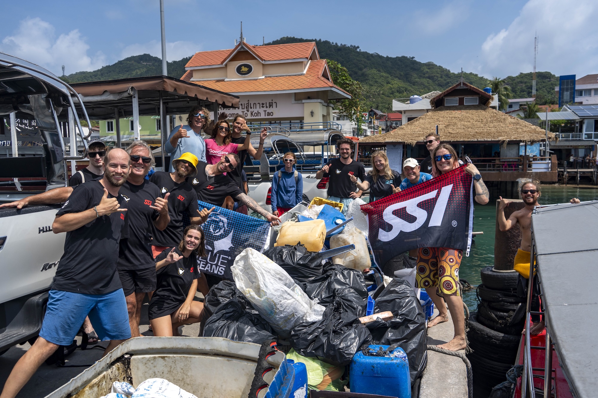 Group of volunteers in Koh Tao celebrate trash cleanup with filled bags and a diving flag, supporting ocean conservation.
