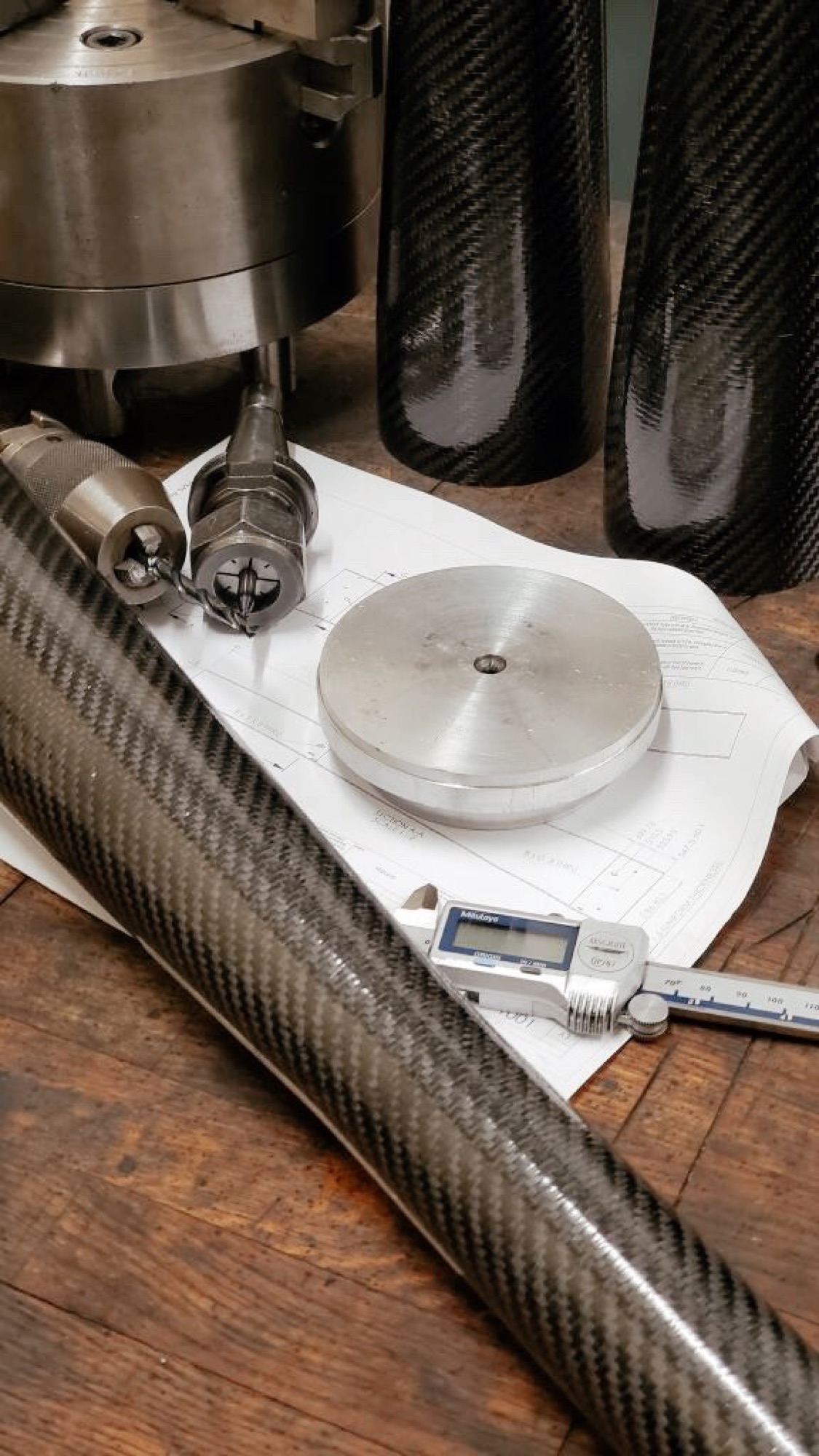 Roll-wrapped carbon fiber tubes with woven surfaces beside calipers, tooling, and a technical drawing.