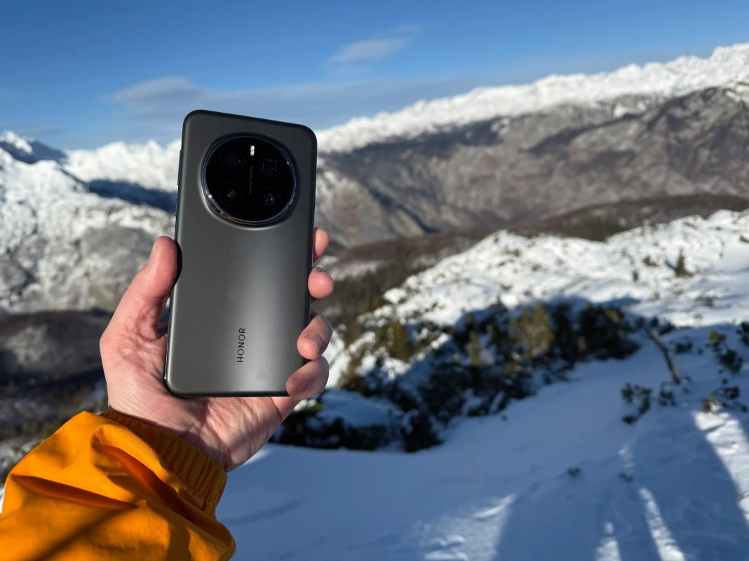 Person holding a smartphone with a mountain landscape in the background, featuring snow-covered peaks and a bright blue sky.