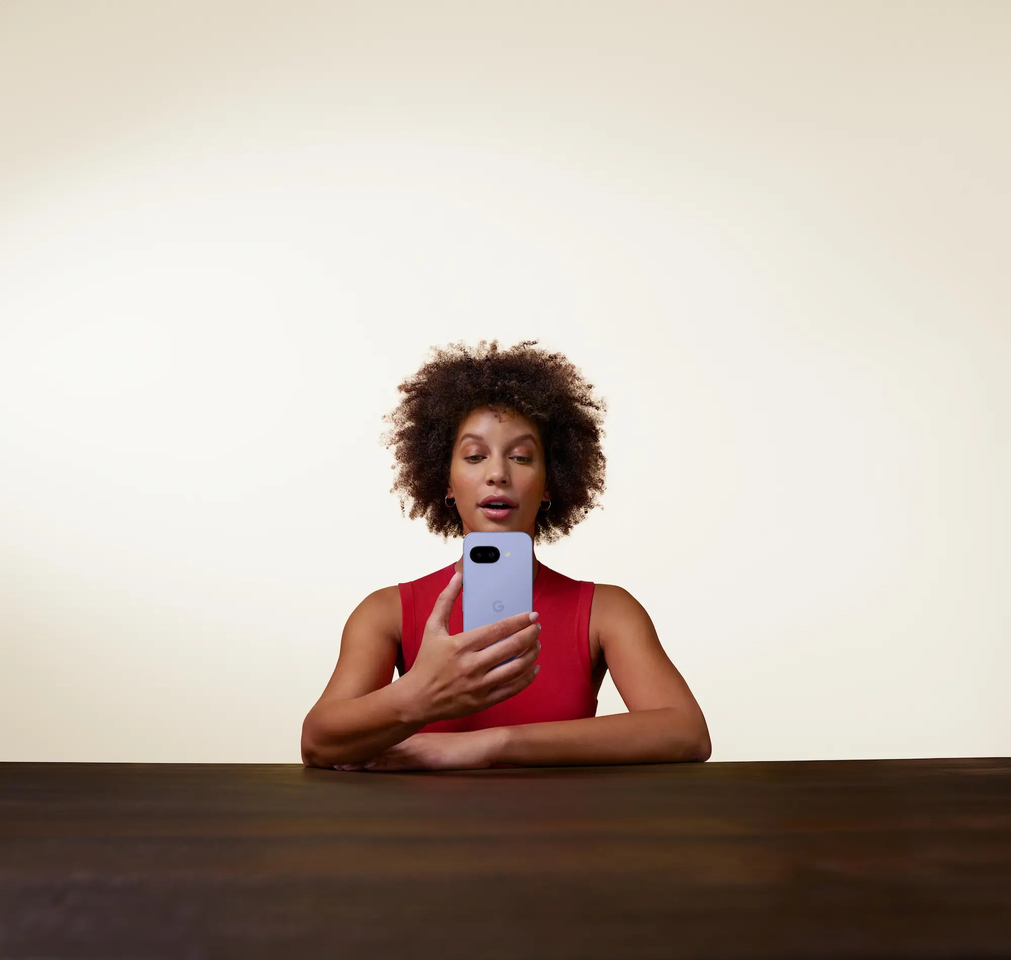 Woman in a red sleeveless top holding a smartphone and looking at the screen, sitting at a dark wooden table against a neutral background.