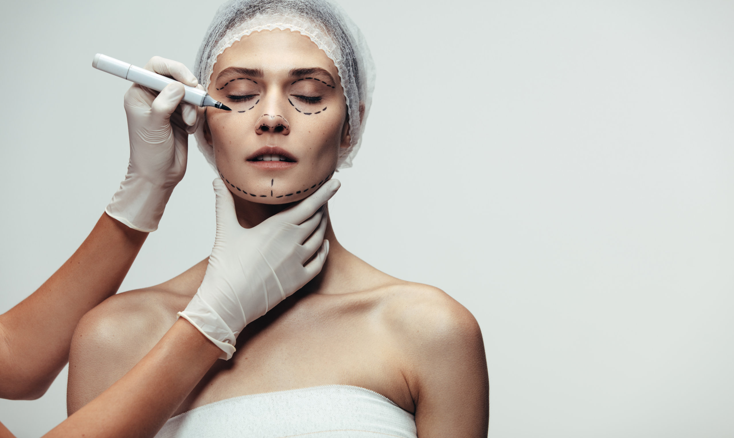 Doctor hands making dotted lines on female face for cosmetic face lift against grey background. Surgeon drawing dotted lines on patient face before surgery.