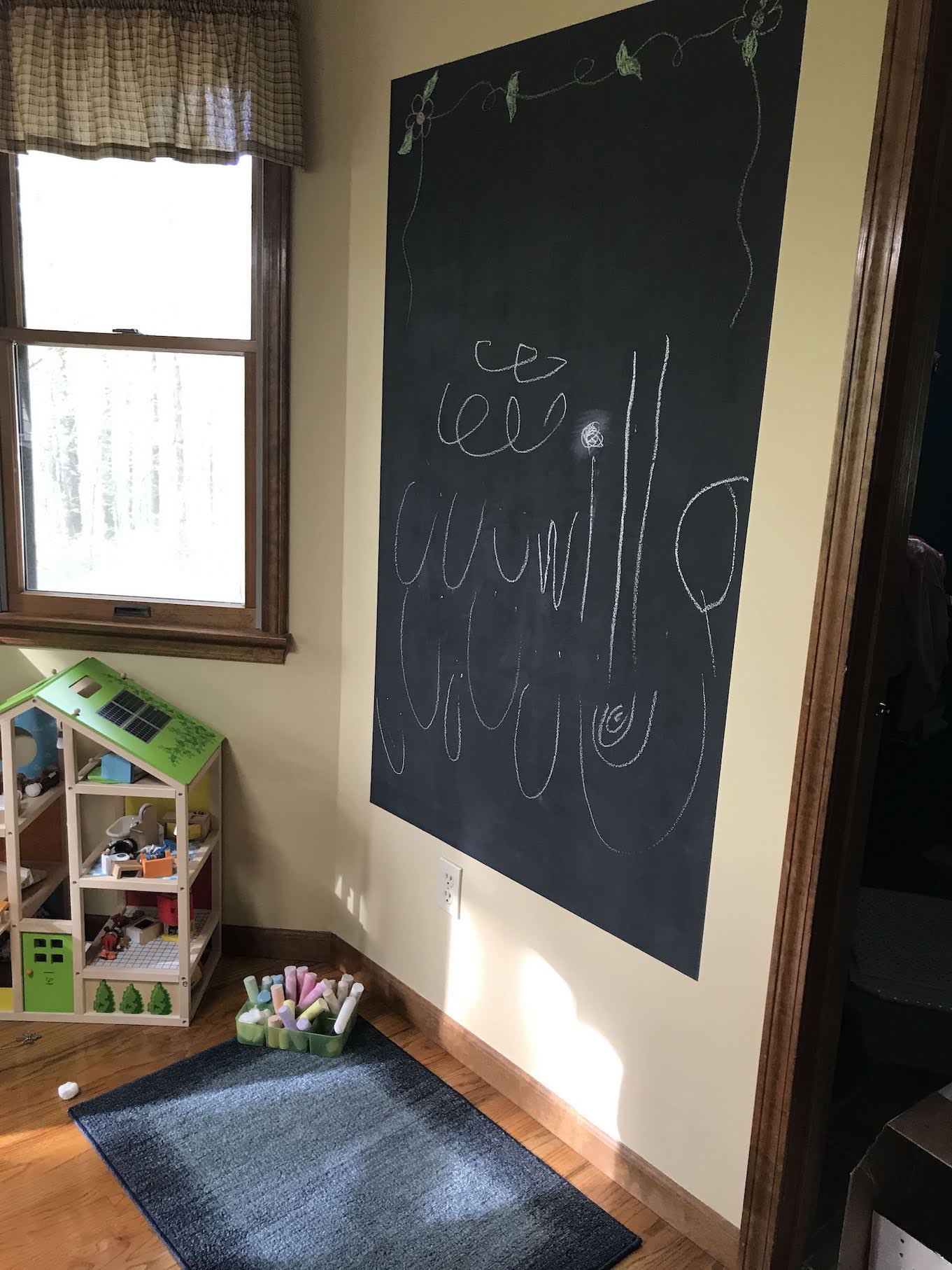 Squiggles and the name 'willa' drawn in white chalk on black DIY chalkboard wall with tray of chalk on floor