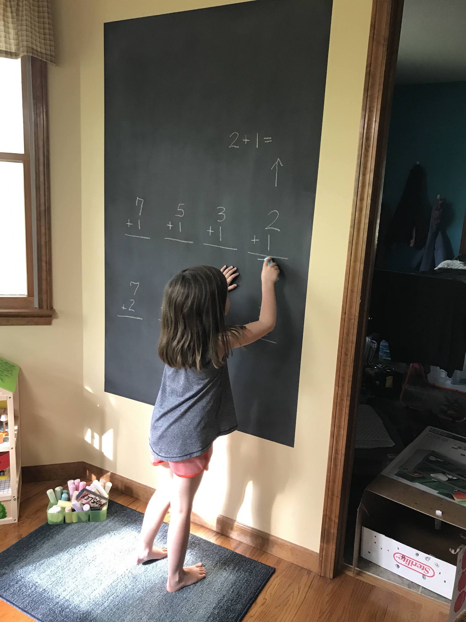 Young girl writing answer to one of five addition math problems written on the black DIY chalkboard wall in white chalk
