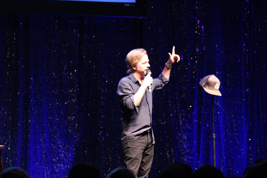 Comedian performing on stage with a hat on a stand