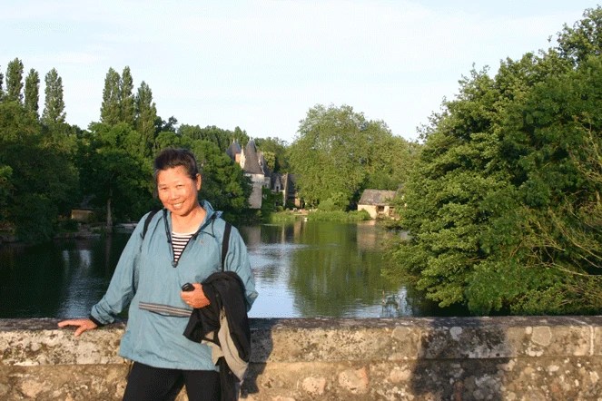 A woman in a blue jacket stands at a stone bridge parapet above a calm river lined with trees, with a turreted house beyond.