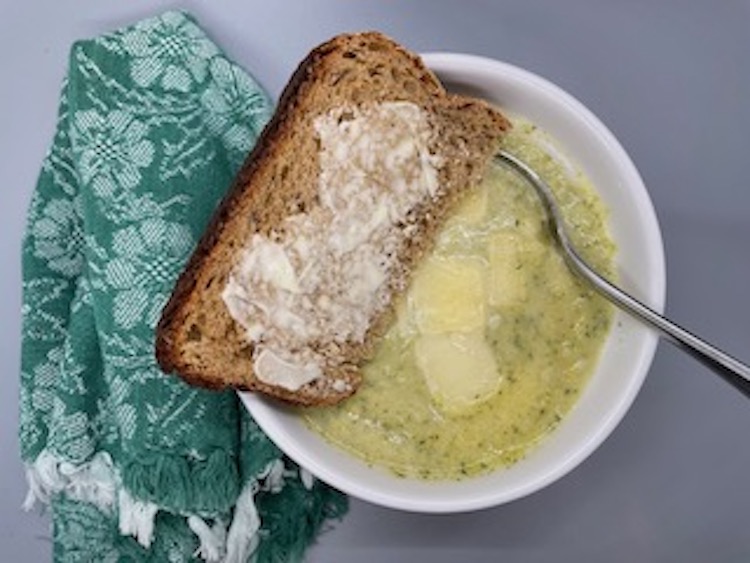 This is a bowl of cream of broccoli soup with a piece of whole grain bread and butter.