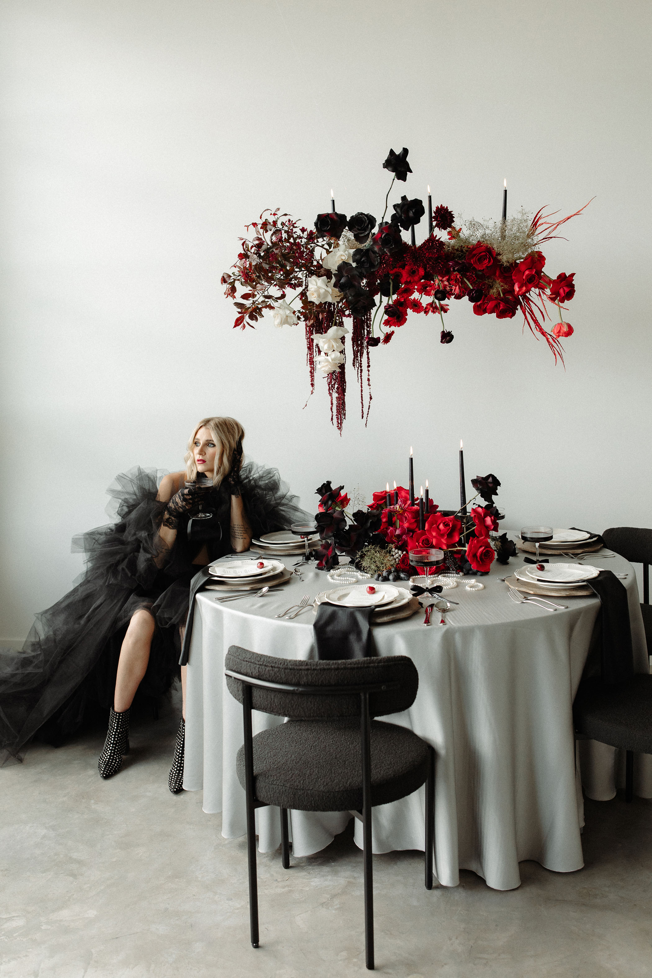 moody red and black Taylor Swift Tortured Poets Department wedding photoshoot with a woman wearing a black tulle dress and sitting at a table with moody hanging florals