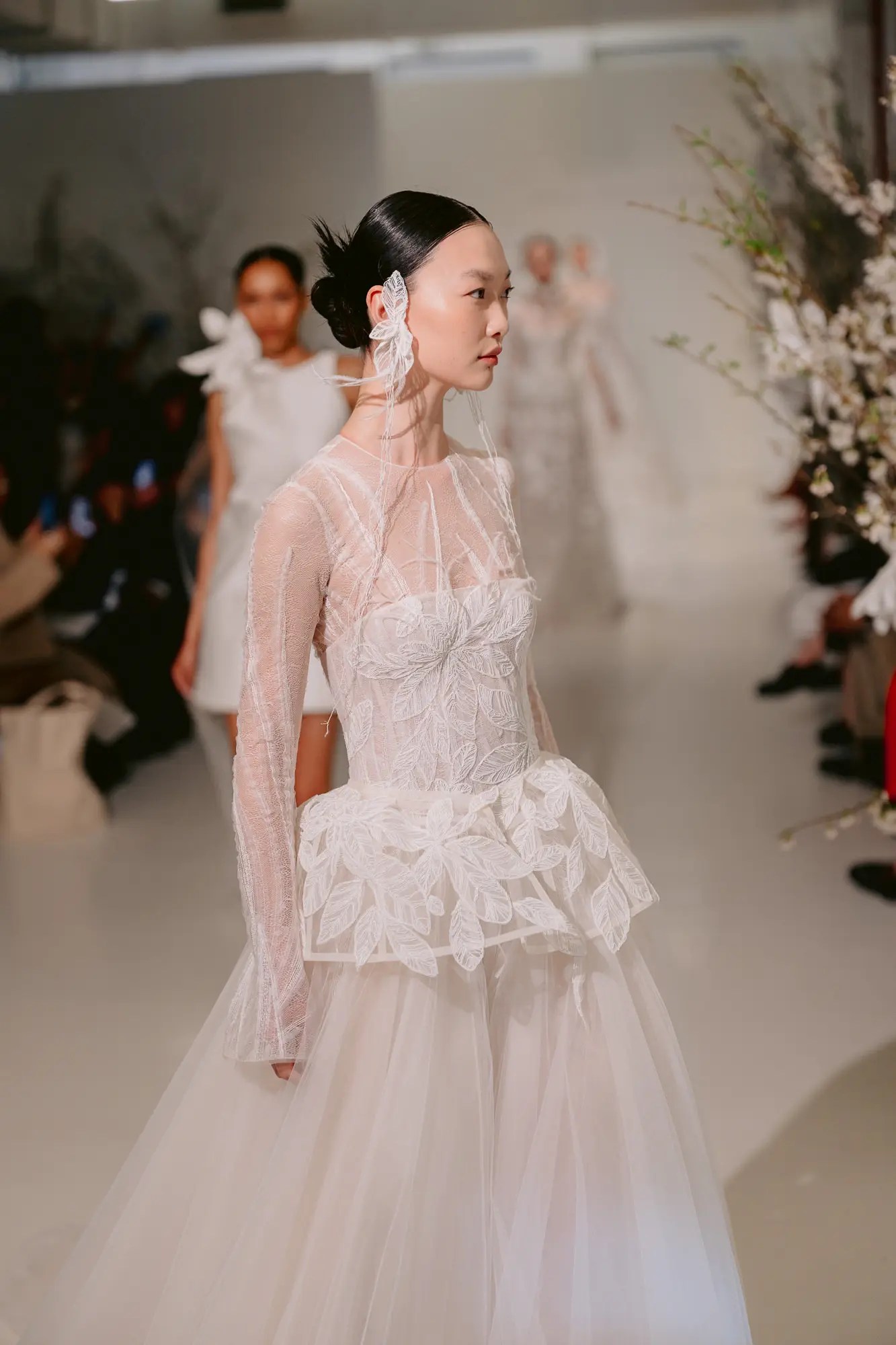 Mira Zwillinger Spring 2027 sheer long-sleeve wedding gown with dimensional leaf appliqué bodice, sculptural peplum waist, and full tulle skirt, styled with a trailing floral ear cuff, shown at New York Bridal Fashion Week.