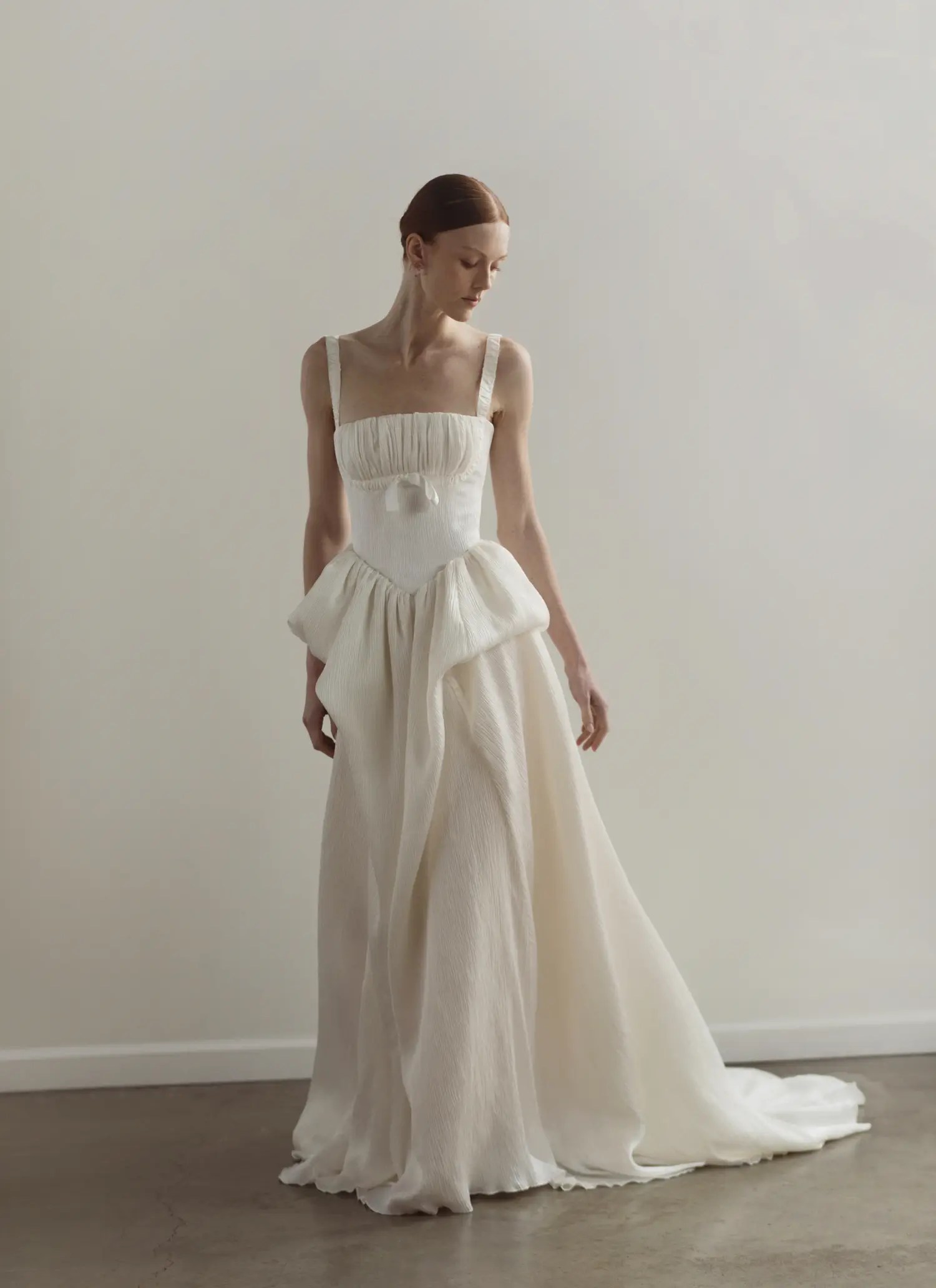 OUMA Spring 2027 ivory square-neck wedding gown with pleated bodice, small bow detail at the waist, sculptural draped peplum, and flowing textured A-line skirt with a sweep train, shown at New York Bridal Fashion Week.