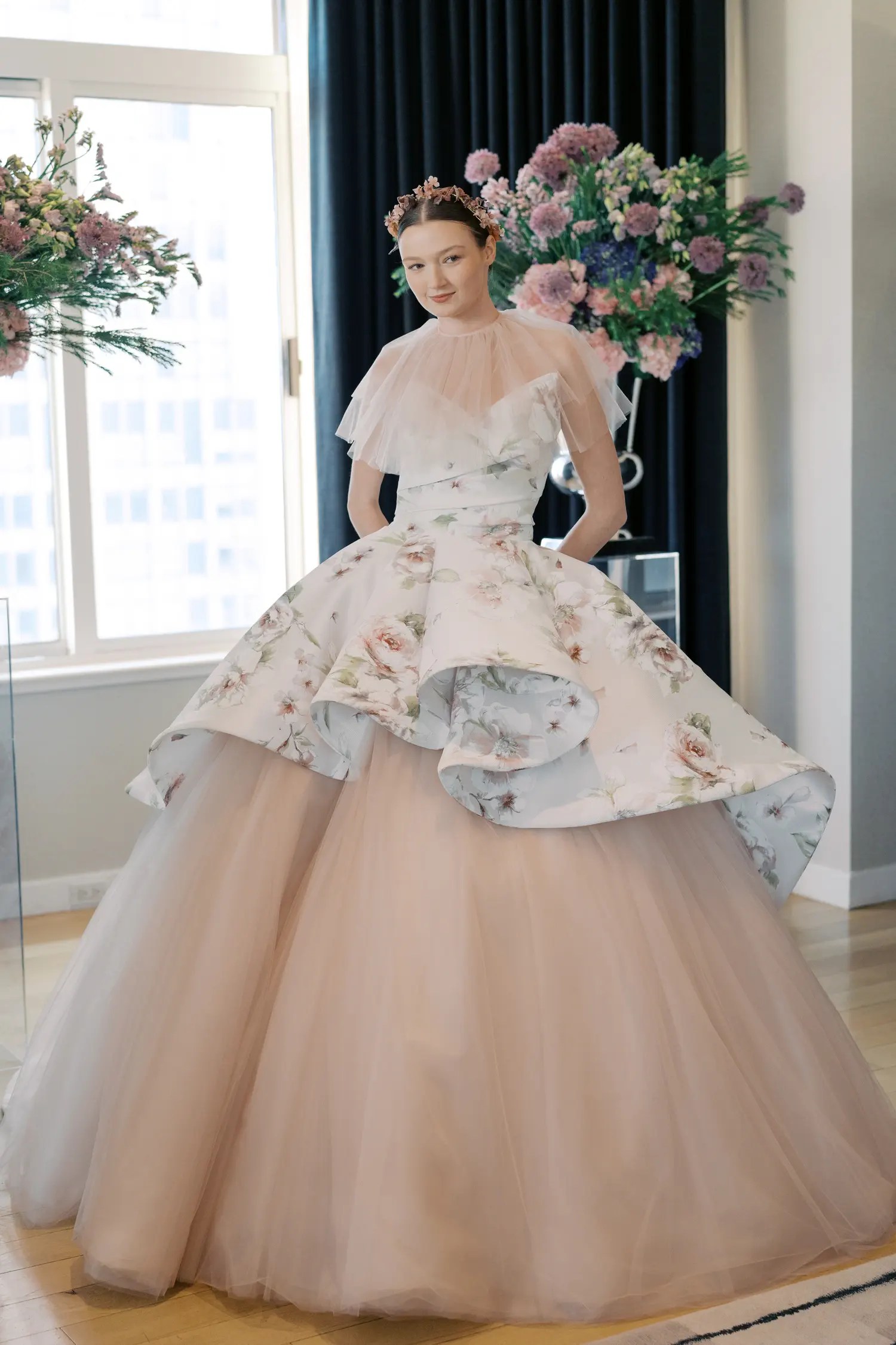 Sareh Nouri Spring 2027 blush ball gown with a sheer ruffled tulle capelet, sculptural floral-printed peplum overskirt in pink and sage, and a full blush tulle skirt, styled with a floral crown headpiece, shown at New York Bridal Fashion Week.