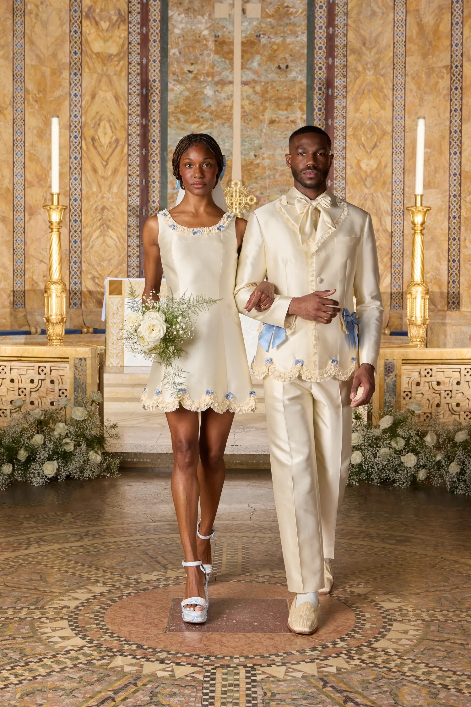 Tanner Fletcher Spring 2027 ivory satin mini dress with blue floral and ruffle trim at the neckline and scalloped hem, paired with a matching ivory satin suit with scalloped trim and blue bow accents, shown at New York Bridal Fashion Week.