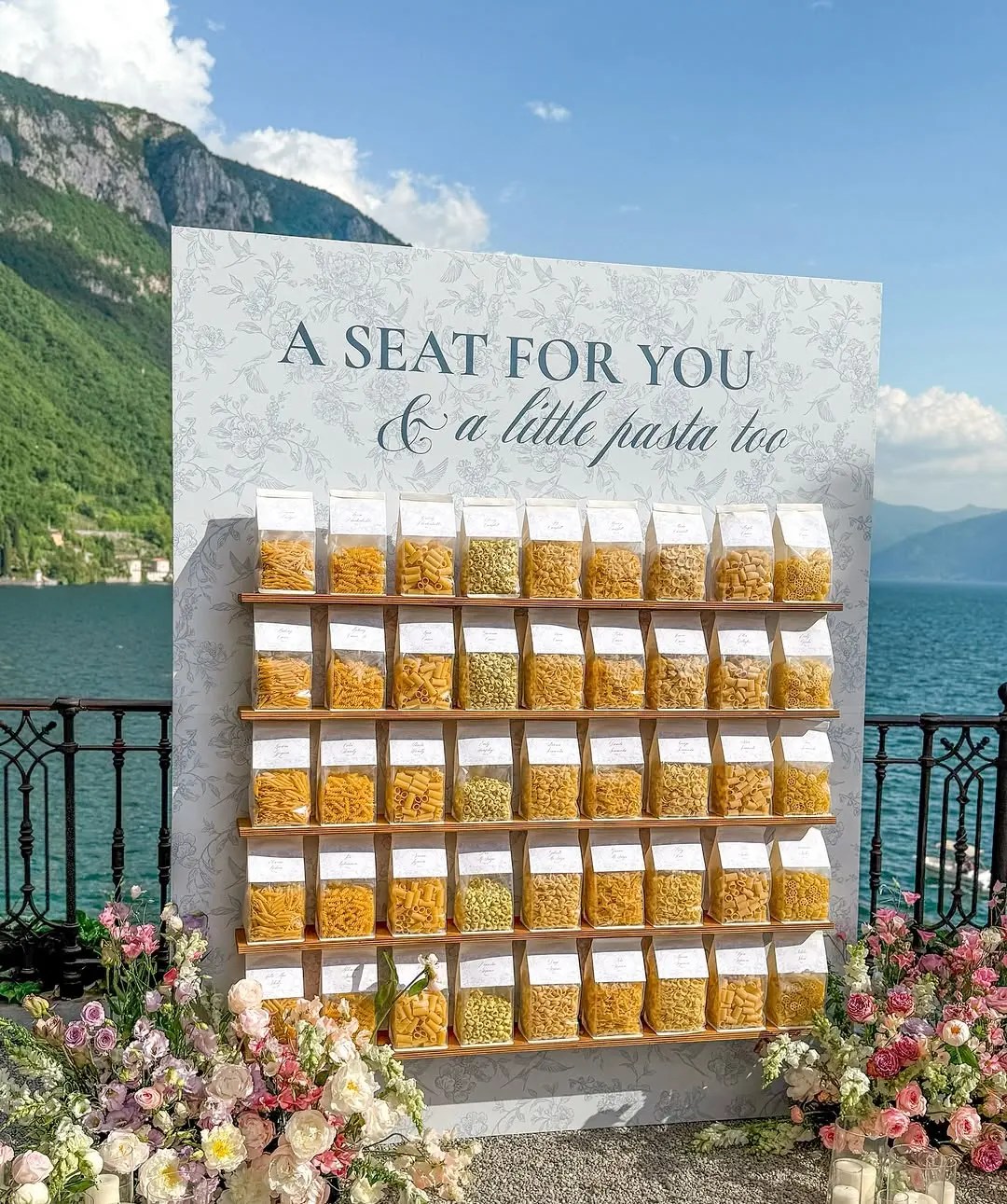 Creative wedding seating chart made with custom pasta packages displayed on a wall at a Lake Como wedding, each labeled with guest names and table assignments, surrounded by floral arrangements overlooking the water.