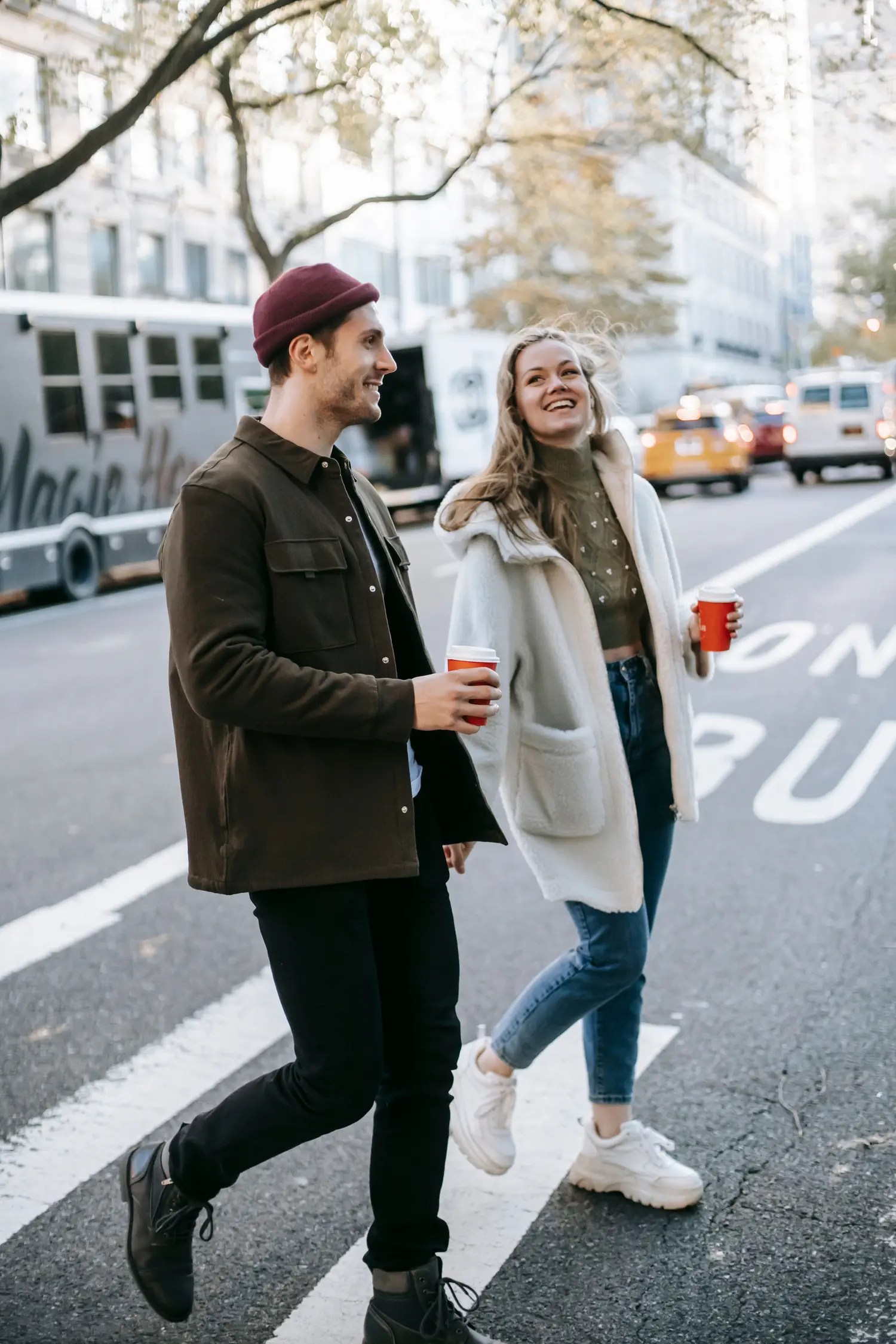 Couple laughing while walking across a city street together with coffees, relaxed and in easy conversation