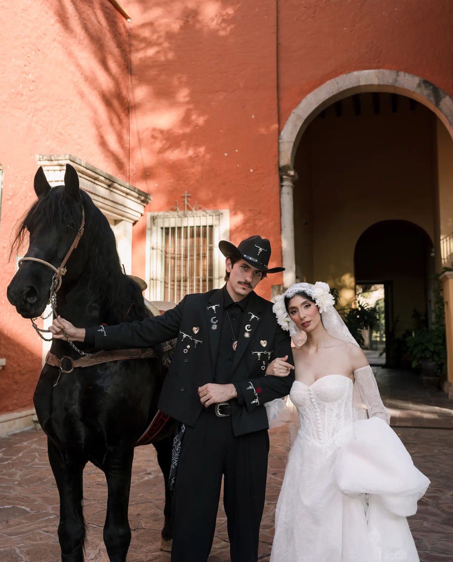 Groom in a black western-style jacket covered in silver longhorn, crescent moon, and heart brooches, standing with his bride and a black horse at a terracotta hacienda