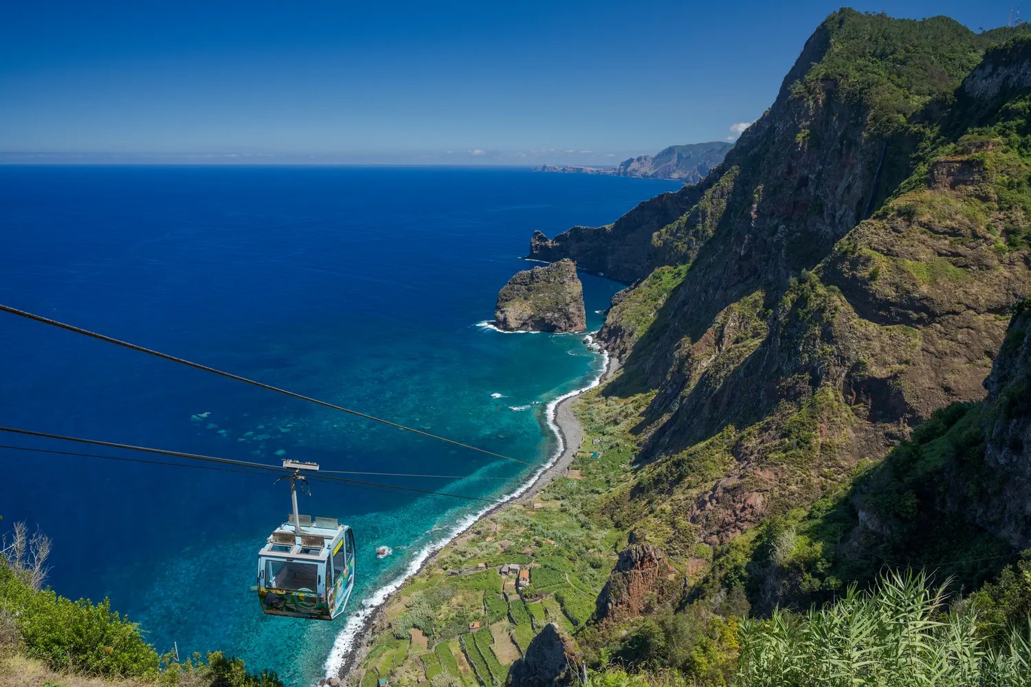 A cable car descending a steep green cliff toward a turquoise cove and pebble beach on the north coast of Madeira