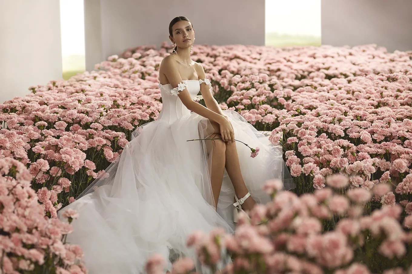 Rosa Clará Couture bride in an off-the-shoulder corset ballgown with floral appliqué sleeves and a full tulle skirt, seated in a field of pink carnations