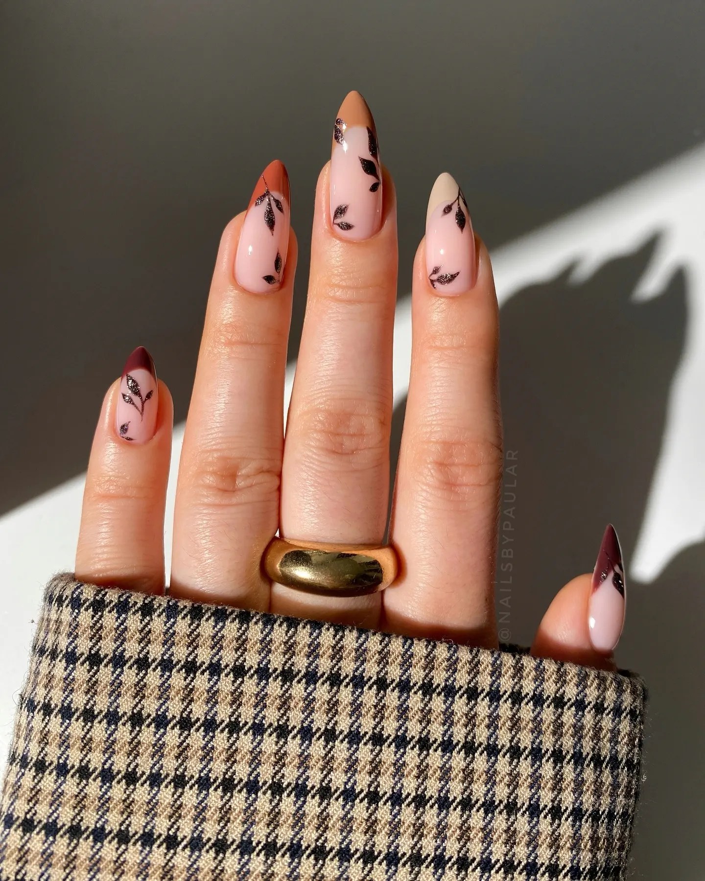 Long almond fall nails with a sheer nude base, tiny black leaf vines and burnt orange and burgundy tips, worn with a chunky gold ring and a houndstooth sleeve.