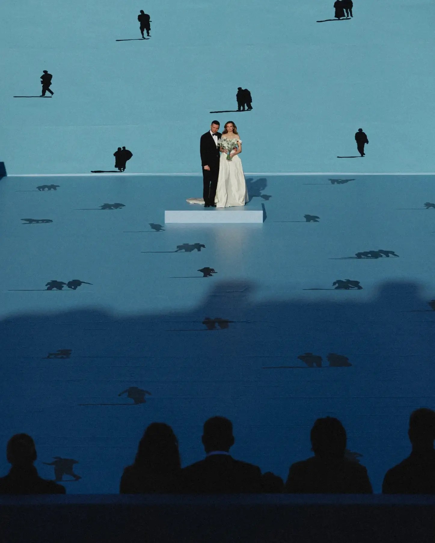 Wedding ceremony on a blue art installation floor with the couple on a white platform, guests silhouetted in the foreground and animal cut-outs casting shadows