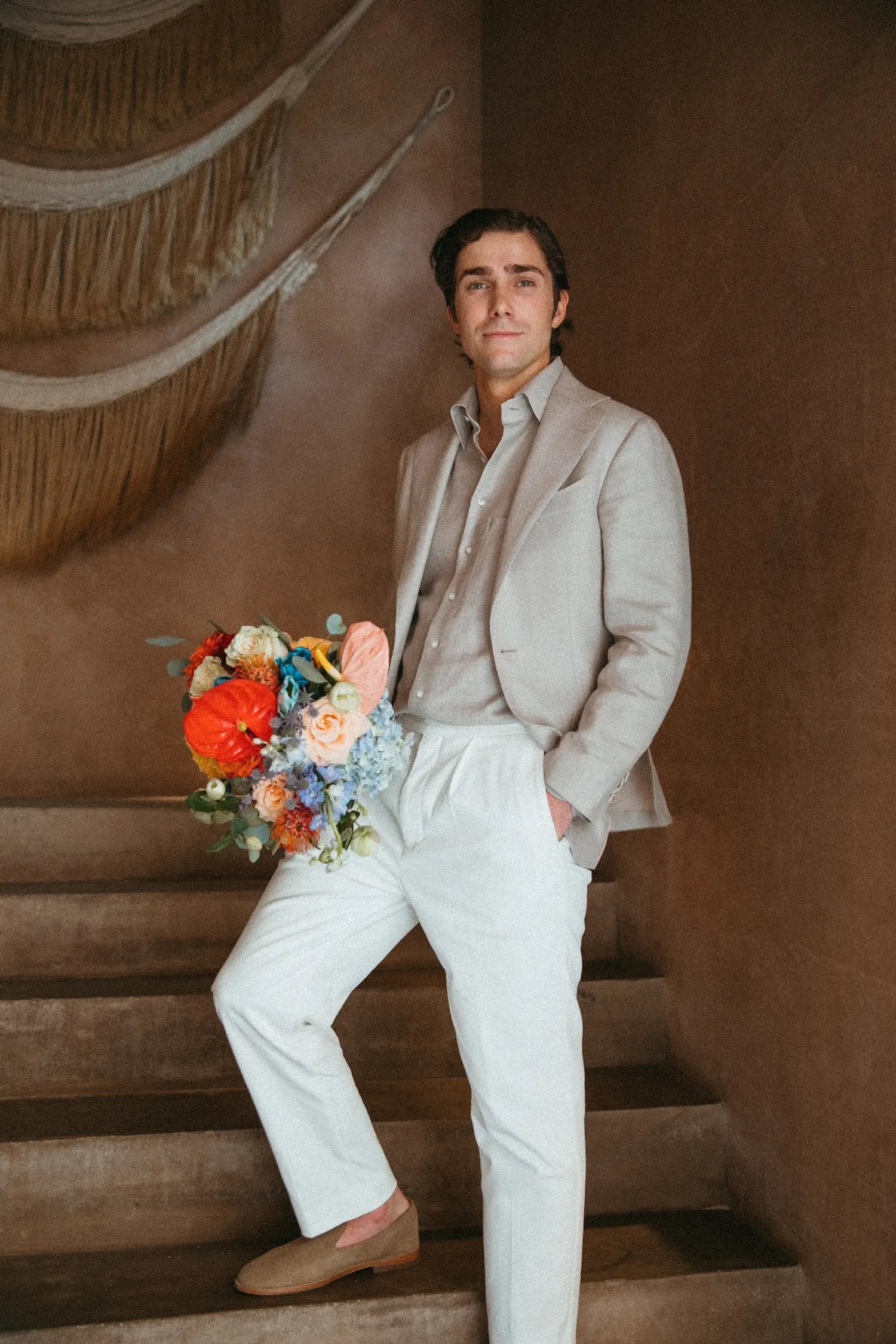 Stephen in a taupe linen suit cradling a bold multicolor bouquet on a terracotta staircase