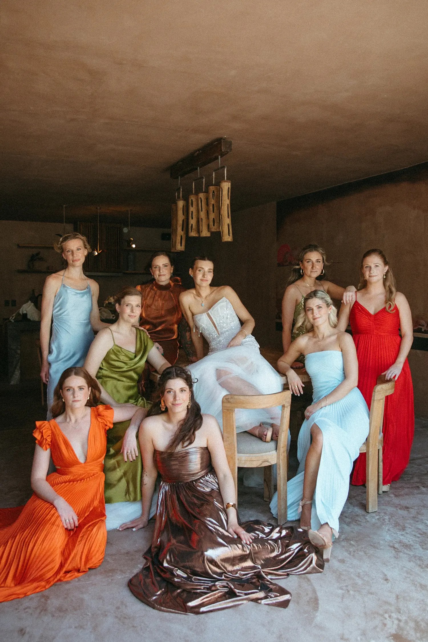 Laura surrounded by nine guests each wearing a different sunset-toned gown in a rustic plastered interior