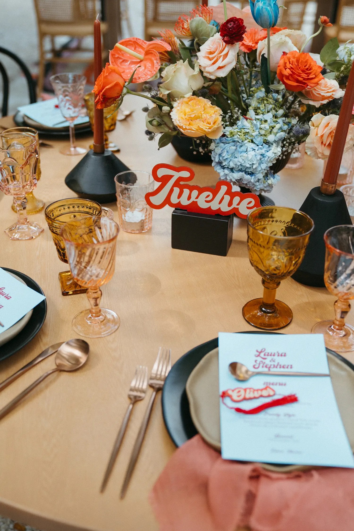 Place setting with a light blue Laura and Stephen menu card in red retro script, amber and pink goblets, and a red acrylic name tag