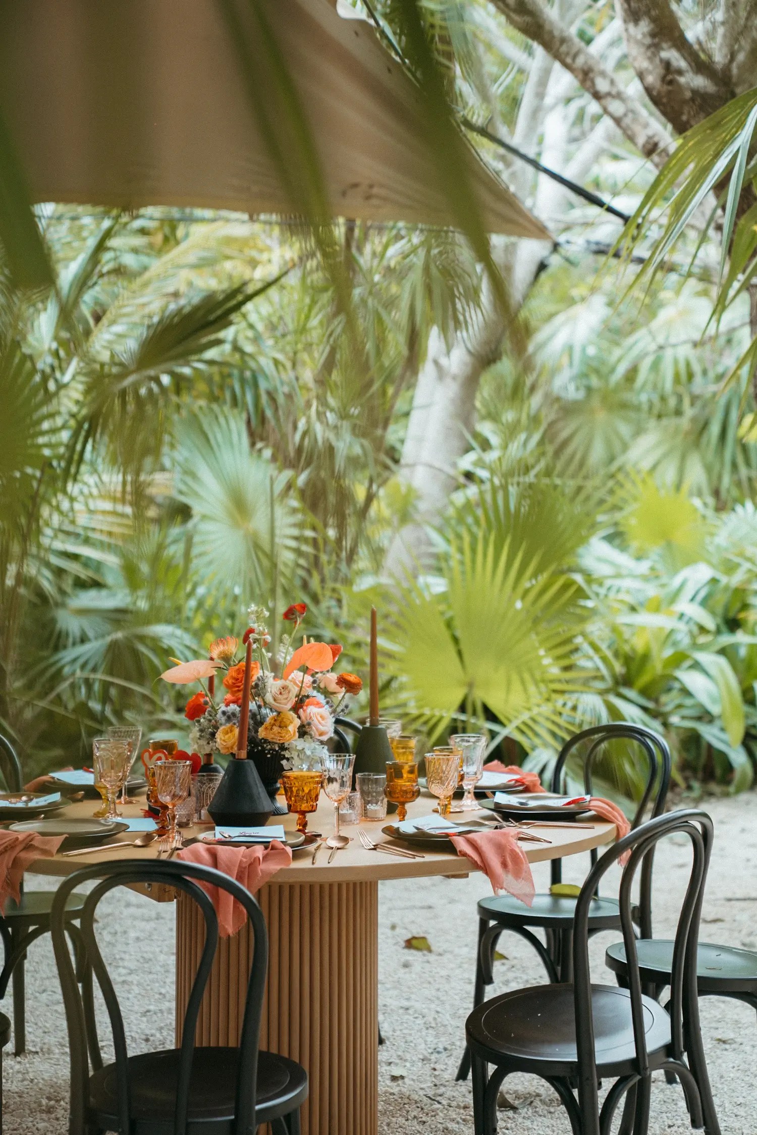 Round wooden reception table with amber glassware, terracotta napkins, and a tropical sunset-toned floral centerpiece