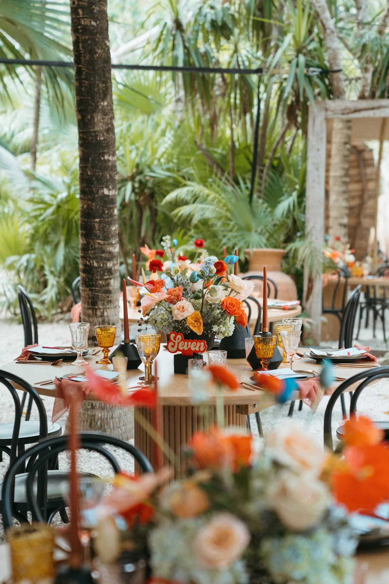 Round reception table labeled 'Seven' with a vivid orange and blue centerpiece and terracotta taper candles in the jungle