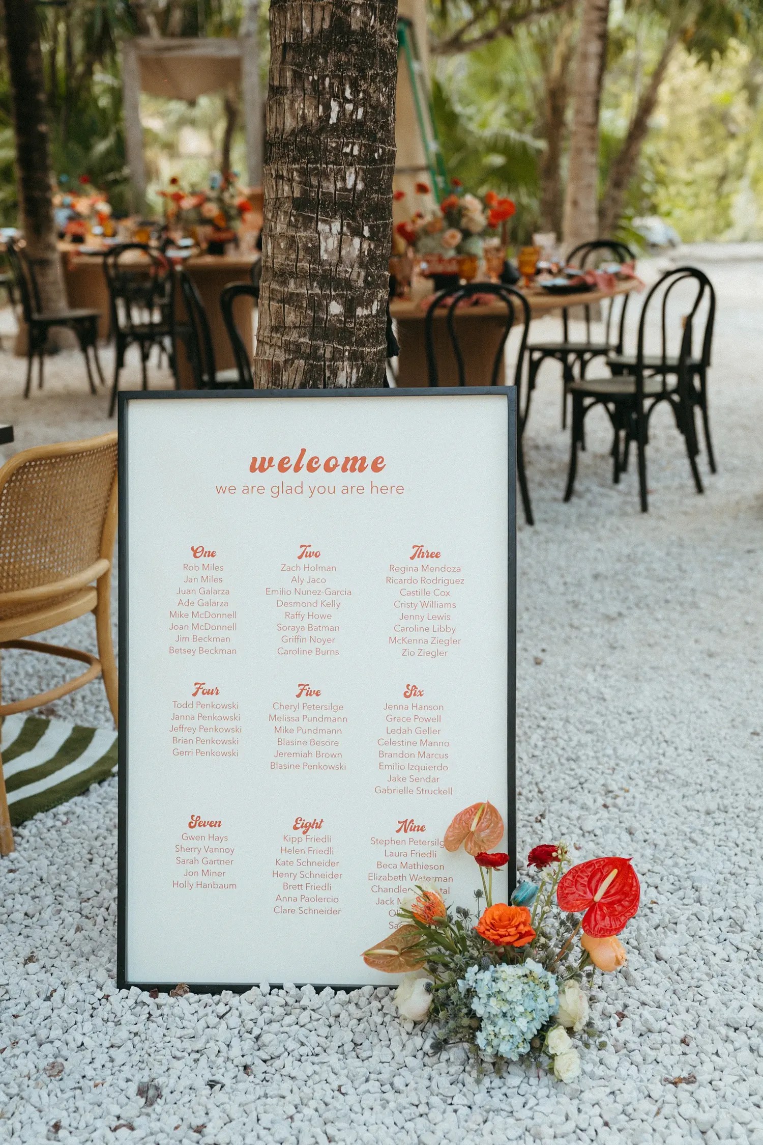 Seating chart sign in retro orange lettering beside a small mixed floral arrangement on white gravel