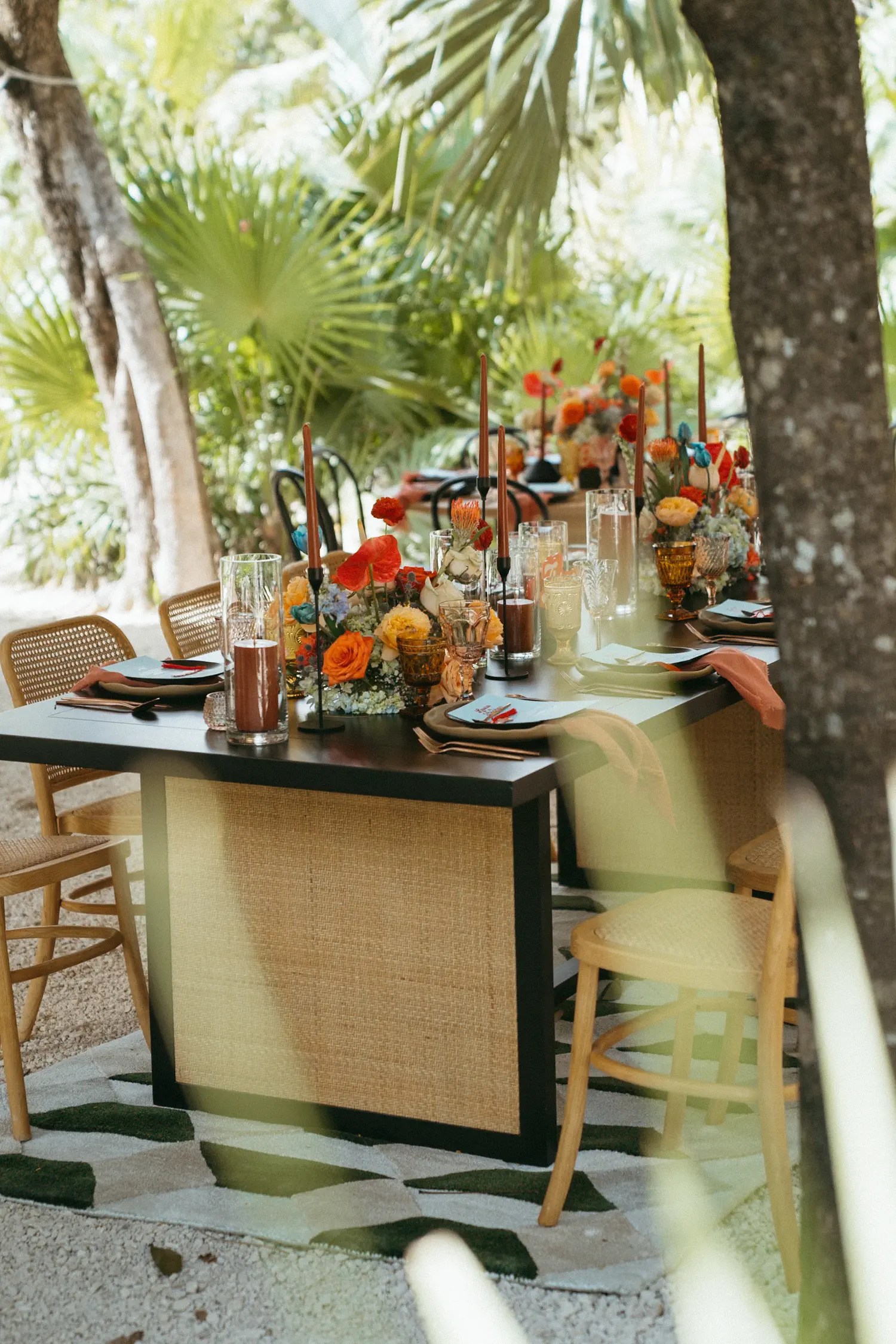 Long reception table with woven side panels, sunset-toned floral centerpieces, and terracotta taper candles under palms