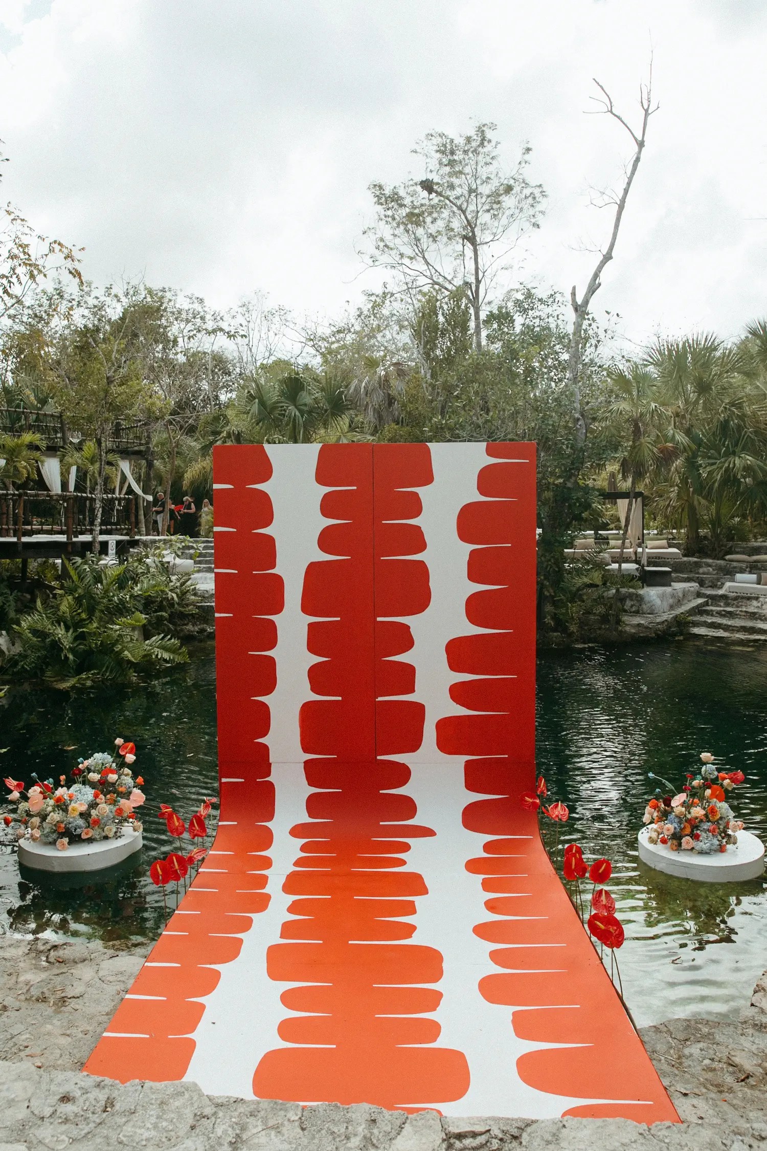 Cenote ceremony altar with a bold red-and-white graphic panel backdrop and matching patterned aisle over the water