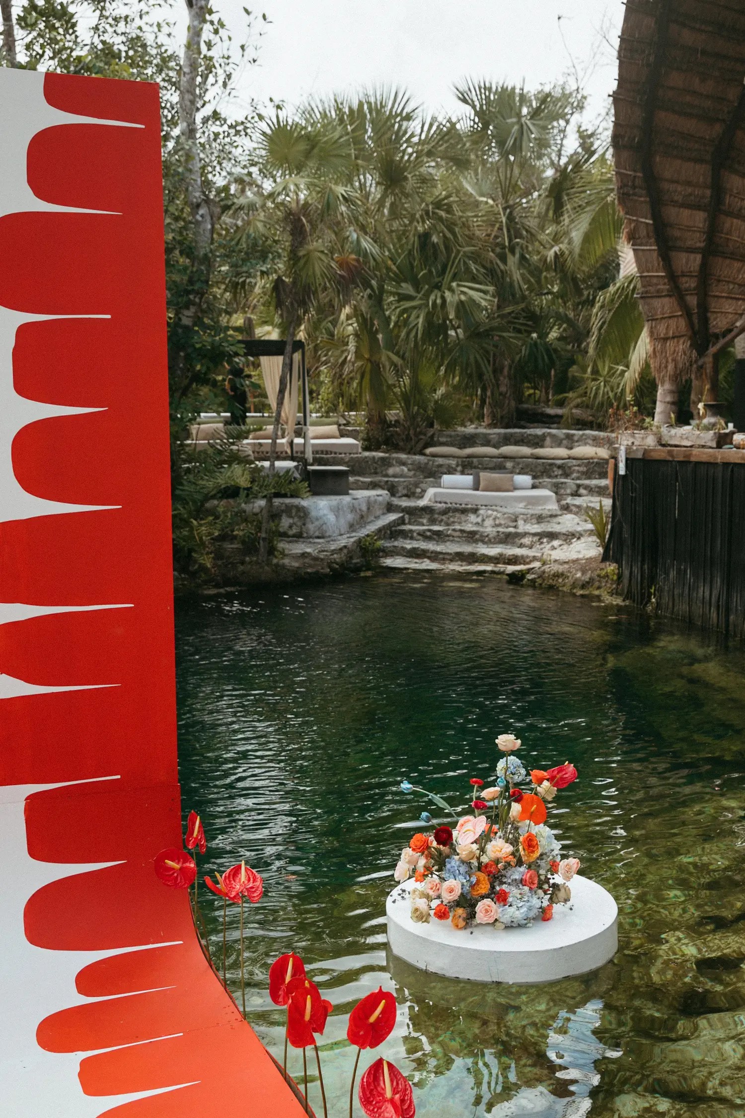 Edge of the red-and-white ceremony panel beside a floating floral pedestal on the teal cenote water