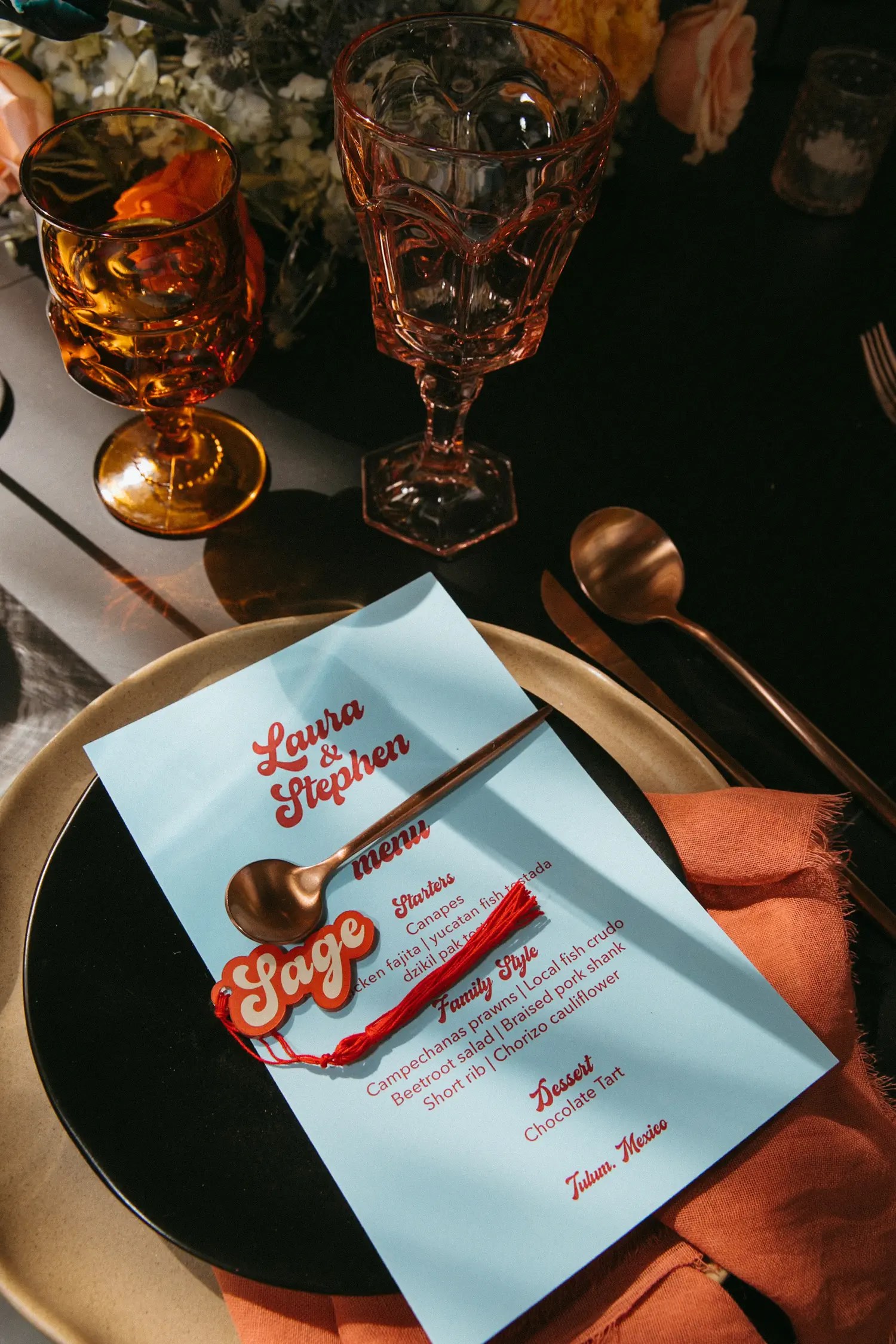 Place setting with a light blue menu card, red 'Sage' name tag with tassel, and gold-rimmed plate on a dark table