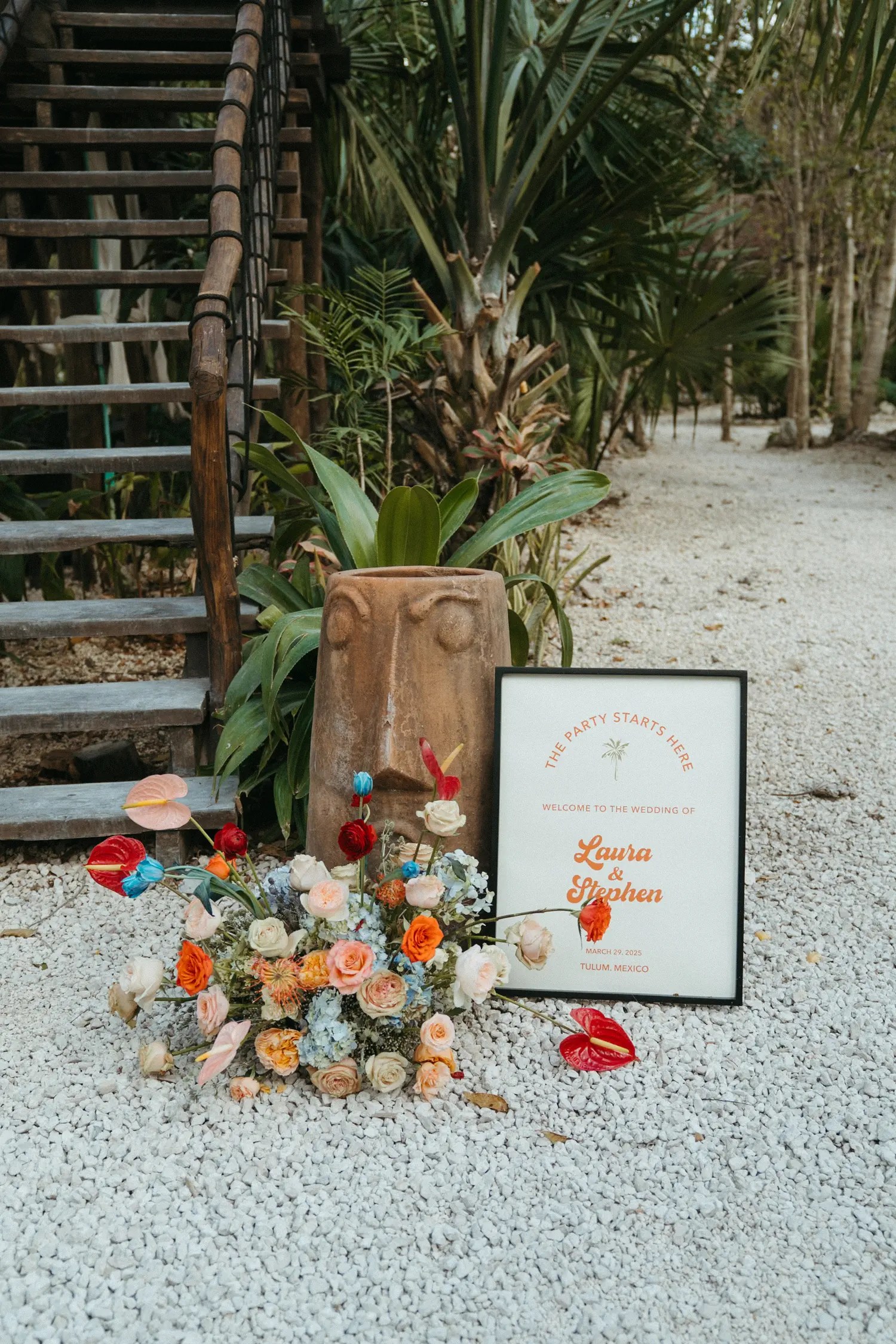 Retro red-and-orange welcome sign for Laura and Stephen beside a clay face vessel and a colorful ground floral arrangement