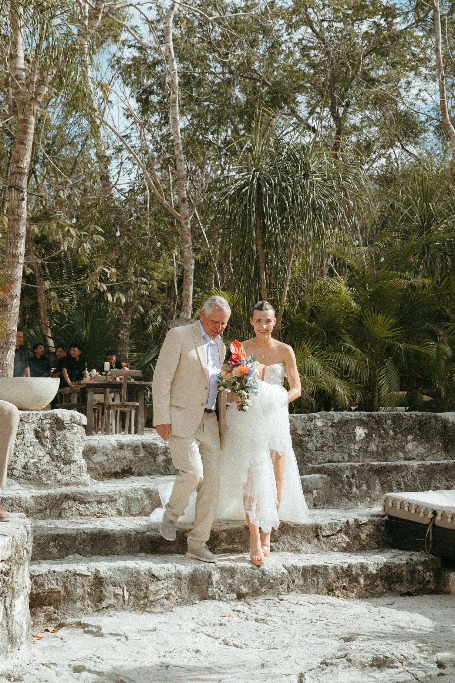 Laura descending rough limestone steps escorted by a man in a cream suit, smiling as she carries her bouquet