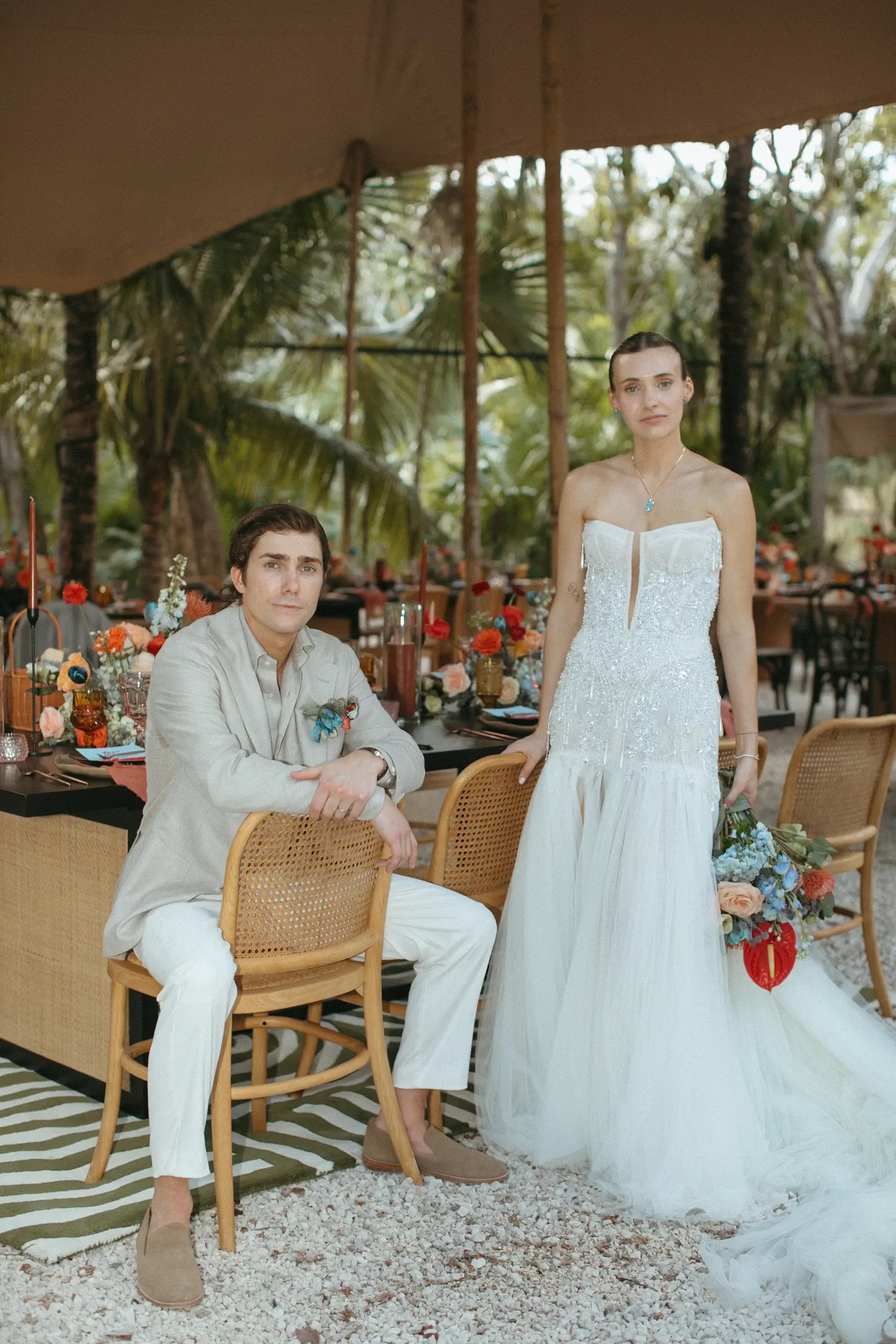 Laura and Stephen at their reception table under a canvas tent, bride in a beaded strapless gown holding a bold multicolor bouquet