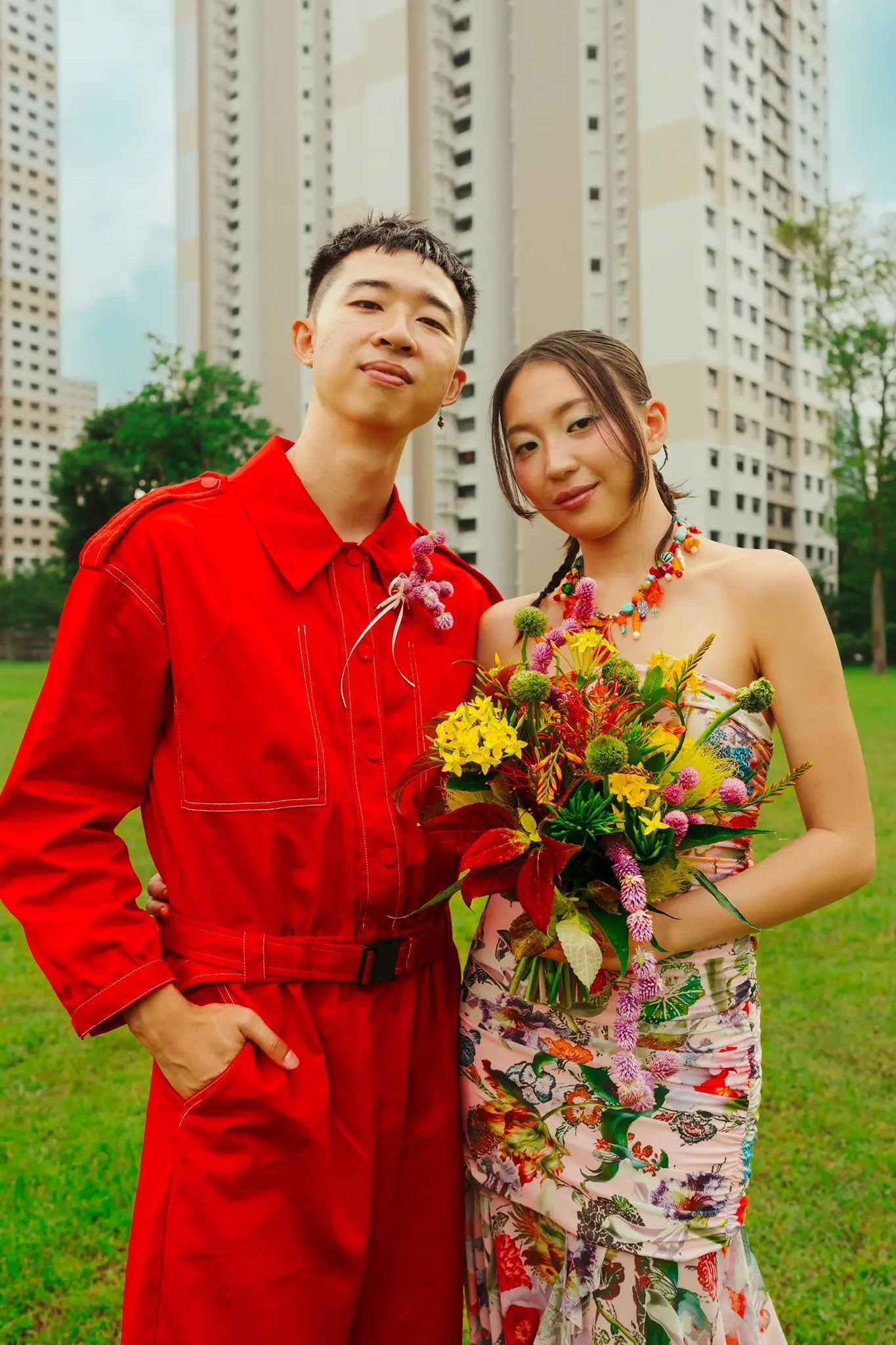 Rio in a red boilersuit with a pink boutonniere and Rachel in a pink floral Desigual dress holding a Meteor Flowers wildflower bouquet on a lawn in front of tall residential towers