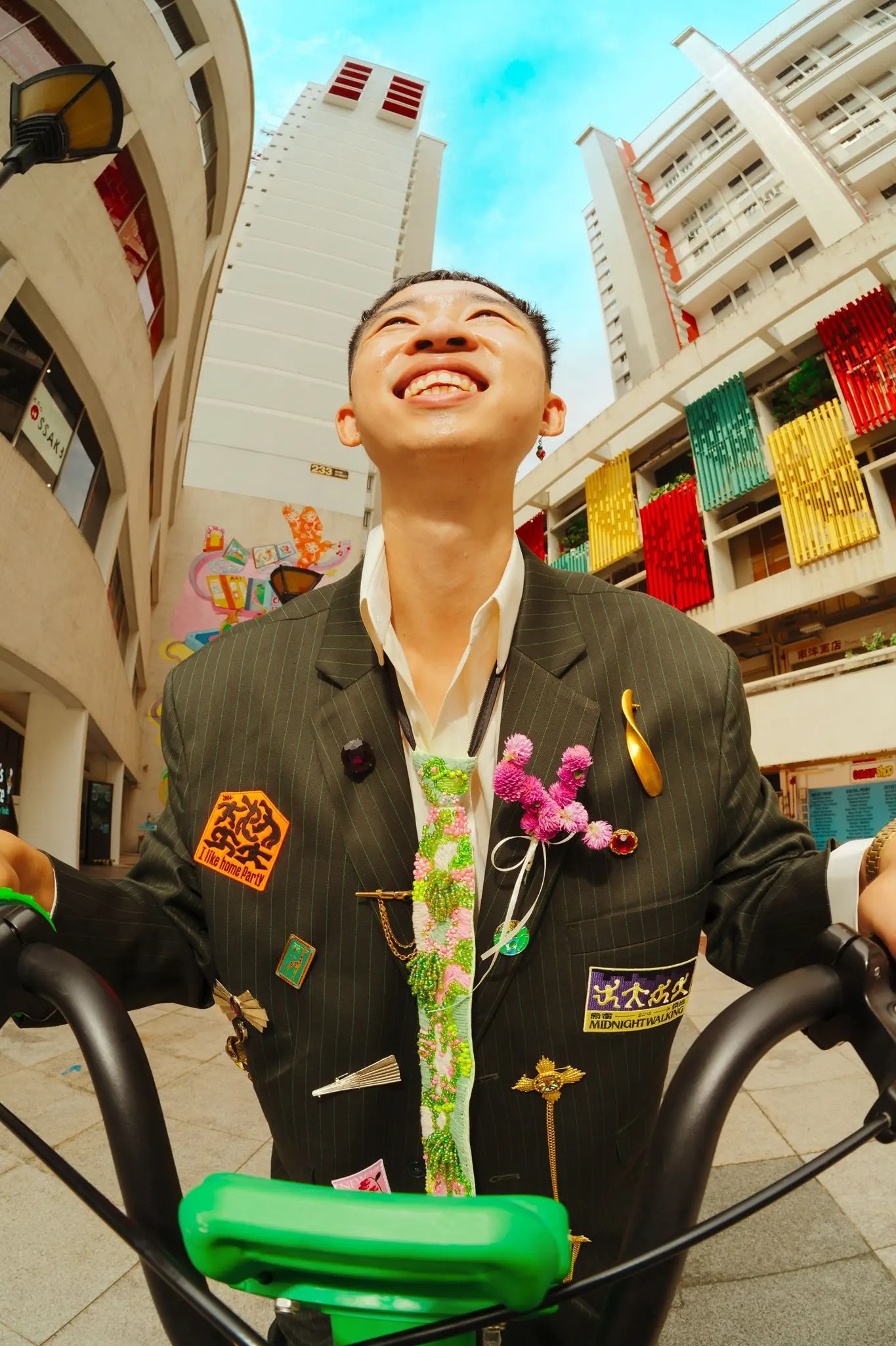 Fish-eye from below of Rio laughing on a green bicycle in a patch-covered pinstripe jacket with pink globe amaranth on the lapel, HDB towers behind